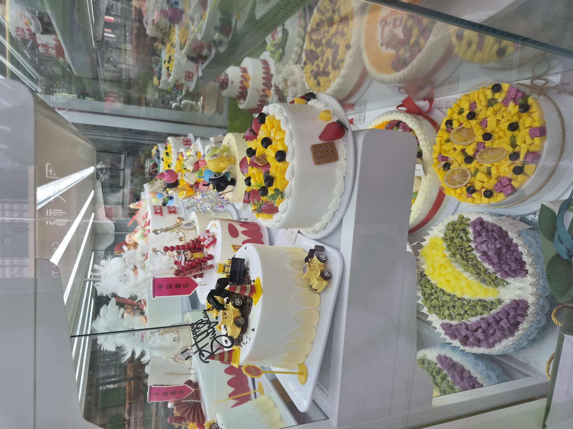 This image shows a display of cakes in a bakery, likely located in Mishan, China, based on the presence of Chinese text on some of the cake decorations. The cakes are arranged on multiple shelves behind a glass enclosure. The foreground features several white-frosted cakes. One cake is decorated with toy construction vehicles, traffic cones, and a "STOP" sign. Another cake is adorned with an assortment of colorful fruit, including mango, blueberries, and strawberries, as well as a waffle-shaped cookie. Further back, a cake with a colorful fruit mosaic design is visible. On the shelves above and to the right, more cakes are displayed, some with fruit toppings and others with decorative frosting patterns. The overall impression is of a well-stocked bakery offering a variety of visually appealing cakes.
