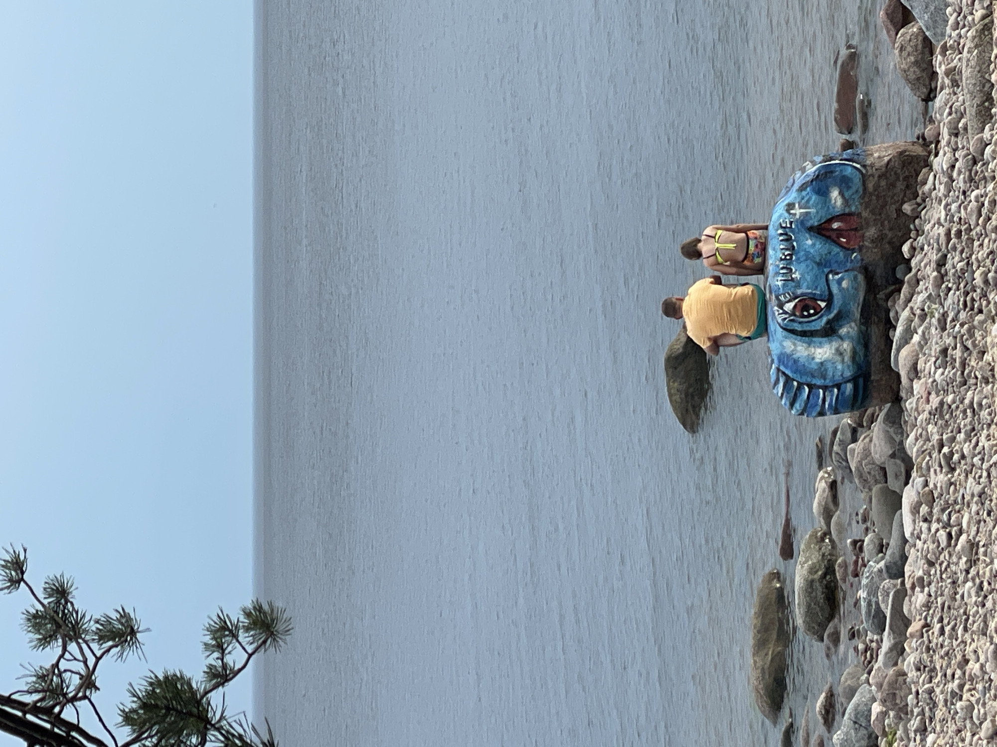 In Luzhki, Russia, two individuals, an adult male and a child, sit on a large, painted rock by the water. The rock is adorned with a blue, cartoonish design of a fish's face with large eyes and a painted cross. The male is wearing a yellow t-shirt and green shorts, while the child is in a bikini. They are facing away from the camera, looking out at a calm, gray body of water under an overcast sky. The shoreline is rocky and scattered with smaller stones. A pine branch is visible in the upper left corner of the frame, extending into the sky.