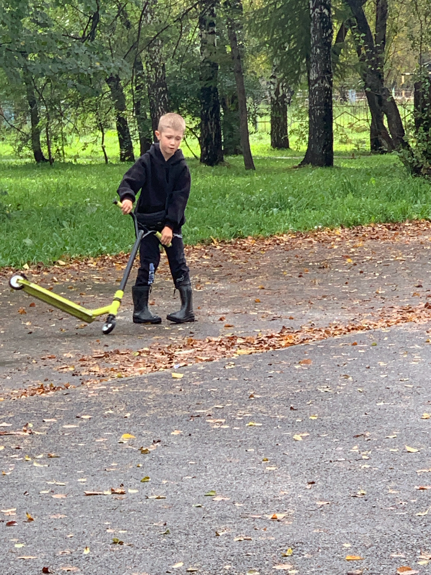 A young boy, appearing to be around 8-10 years old with light-colored hair, stands on an asphalt path in a park. He is wearing a black hooded sweatshirt, black pants, and black rubber boots. The boy is holding the handlebars of a lime green and black scooter, which is positioned in front of him, slightly to his left. The scooter's front wheel is turned to the left. The path is covered with scattered brown and yellow leaves, indicating an autumn setting. To the left of the path is a grassy area, and behind that, a dense cluster of trees with green foliage and some bare branches. The background suggests a natural, wooded park environment within the city of Prokopyevsk, Russia. The overall scene depicts a moment of leisure as the boy prepares to ride his scooter.