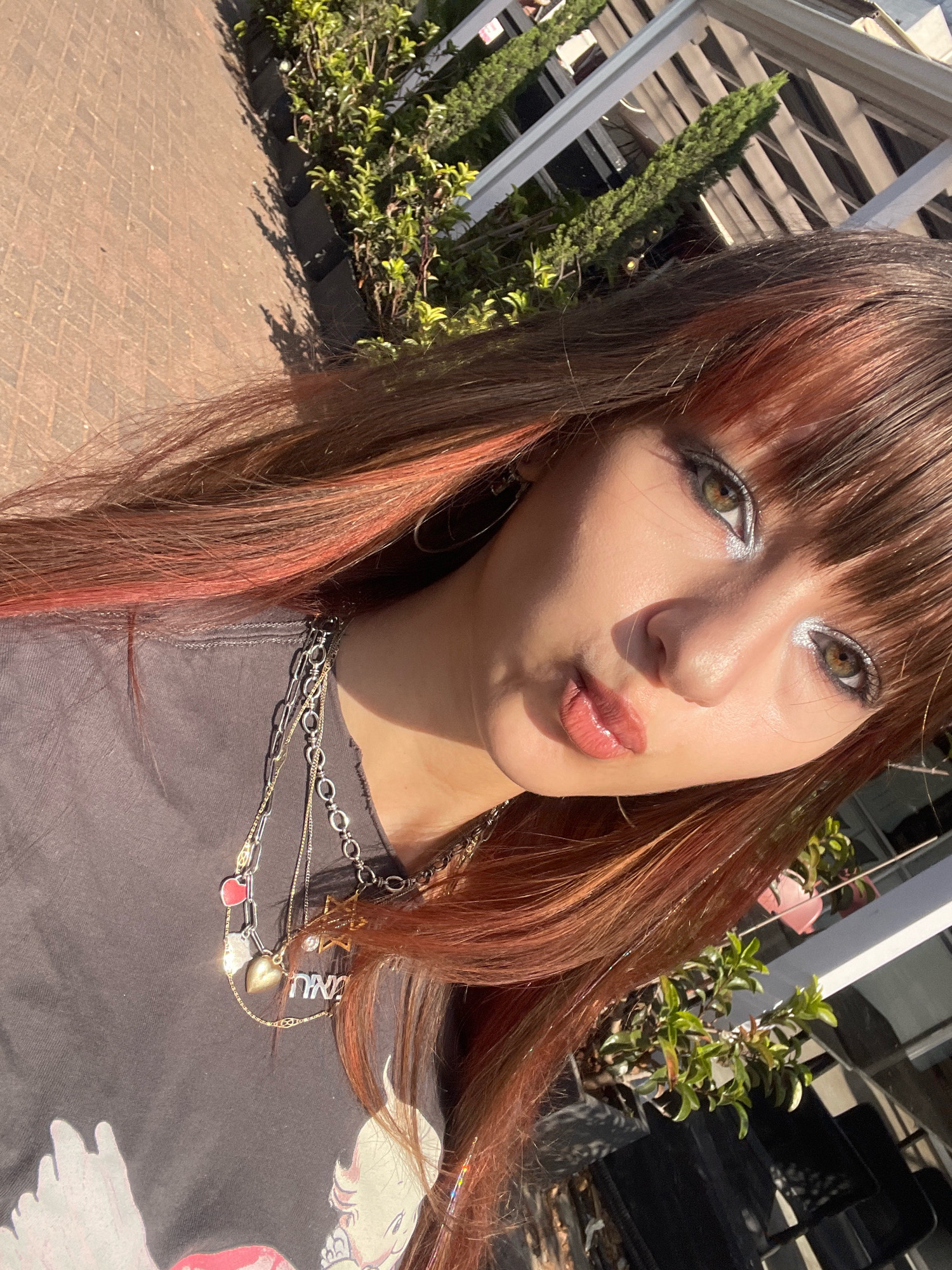 The image is a close-up, selfie-style shot of a young woman outdoors, likely in Raanana, Israel, given the context. The woman has long, auburn-streaked brown hair and is wearing a dark gray t-shirt. She has on noticeable makeup, including silver eyeshadow and lip gloss. She is adorned with multiple layered necklaces, one of which features heart and star charms, and another with a pendant bearing Hebrew letters.
The background suggests an outdoor setting with elements of urban landscaping. To the left, a reddish-brown brick pathway is visible, possibly a sidewalk or patio. To the right and behind her, there is lush green foliage, suggesting plants or bushes, with a white wooden or metal structure that appears to be a fence or pergola casting shadows. Sunlight is prominent, creating highlights on her face and hair, and casting distinct shadows. The overall scene conveys a casual, daytime setting in what could be a park, a garden, or a residential area with outdoor decor.