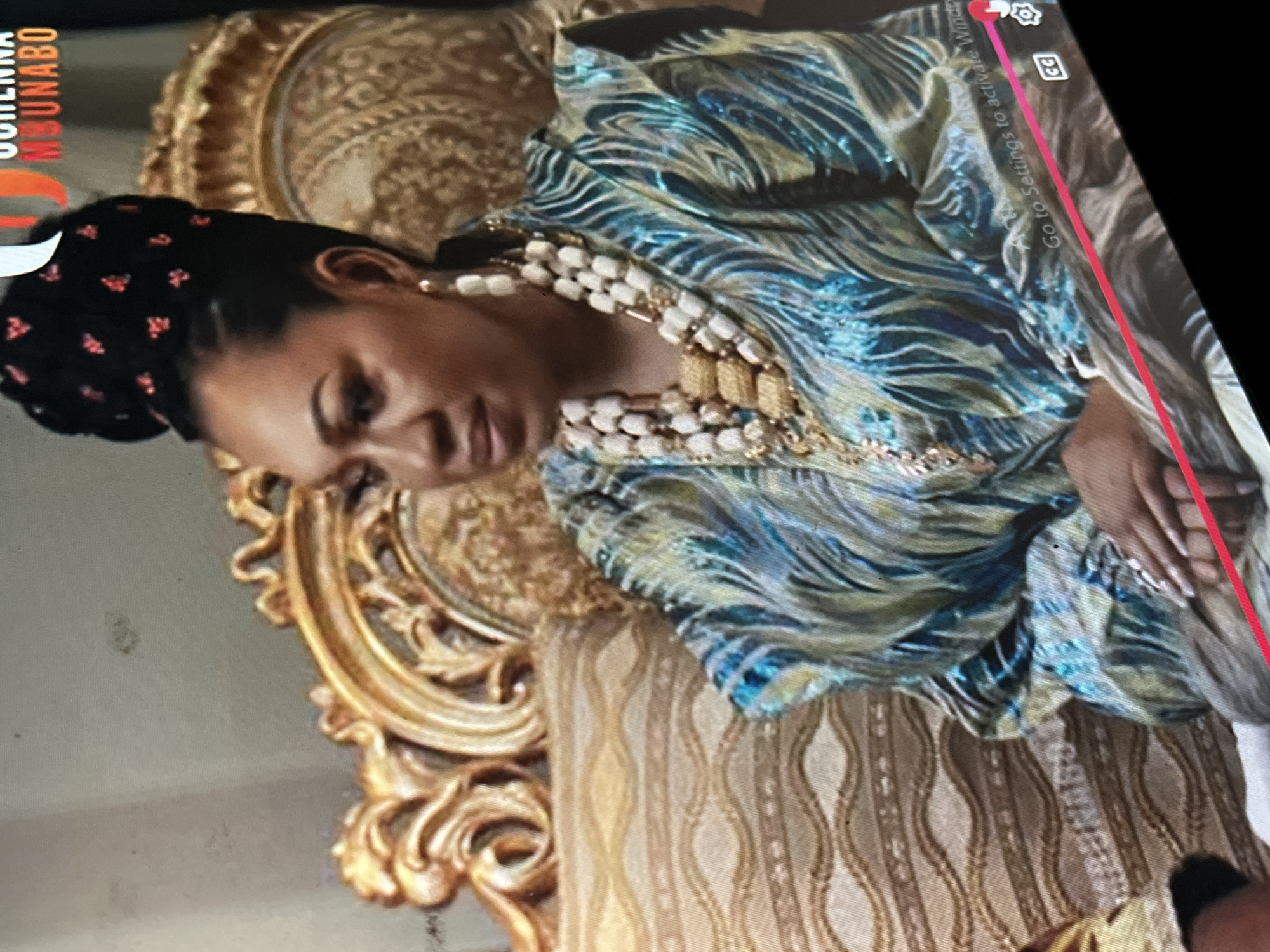The media file captures a close-up, rotated shot of a woman in an indoor setting, likely in Abuja, Nigeria, given the provided context. The image appears to be a photograph of a screen displaying a video or picture.
The main subject is a woman with dark skin and dark hair styled in an intricate updo, adorned with numerous small, red, possibly floral or geometric embellishments. She is wearing a striking garment with a wavy, iridescent pattern in shades of blue, green, and gold. Her attire is accessorized with an elaborate, multi-layered necklace featuring prominent white beads and gold rectangular elements. She also wears pearl-like earrings. Her head is tilted downwards, and she is looking away from the camera, giving a contemplative or demure impression. Her left hand, with long, manicured nails, is visible resting on an unseen surface.
She is seated in an opulent, gold-colored chair with highly decorative, carved wooden frames and upholstered in a patterned fabric featuring vertical wavy lines in beige and gold tones. The ornate back and armrest of the chair are visible, highlighting its elaborate design.
The image contains several screen overlays:
- Vertically on the left, partially obscured, is the text "UCHENNA MBUNABO" in red and white.
- On the upper right, a settings gear icon and a "CC" (closed captions) icon are visible. Below these, a partially obscured message reads "ACT Go to Settings to activate Window", indicative of a Windows operating system watermark.
- A prominent red vertical line runs down the right side of the screen, possibly indicating a progress bar or a UI element.
The lighting appears bright, illuminating the intricate details of her attire and the chair. The overall scene conveys a sense of elegance and cultural richness.