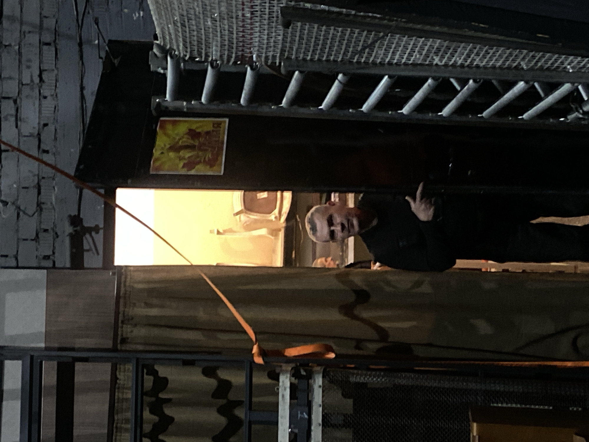The image shows a man with short gray hair, wearing a dark-colored shirt or jacket, partially visible through an opening in a dark, boxy structure. The man's face is in view, and he is pointing upwards with his right index finger. Above the structure is a metal grate or mesh. To the left is a partially visible brick wall. Below, there appears to be some kind of stage or platform structure with draped material. An orange strap hangs down from the upper structure. A sticker or small poster is attached to the boxy structure. No additional information regarding the city of Заволжье, Russia, is discernible from the image.