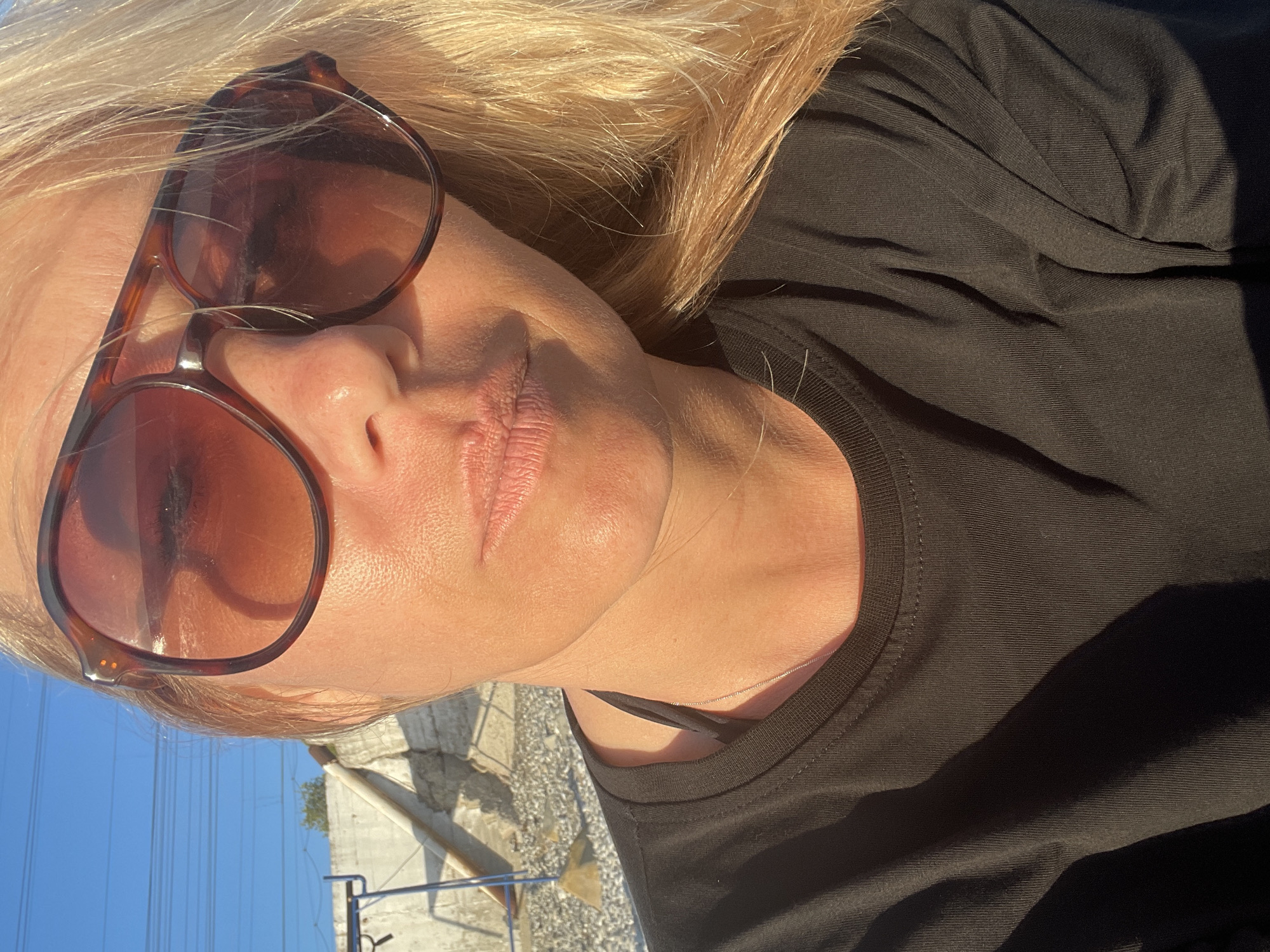 The image is a close-up selfie of a woman outdoors, likely on a sunny day. She has blonde hair and is wearing large, tortoise-shell sunglasses and a black t-shirt. Her skin appears tanned. In the background, a concrete structure and some gravel are visible, suggesting a potentially urban or coastal environment. The lighting indicates it is either morning or late afternoon.
