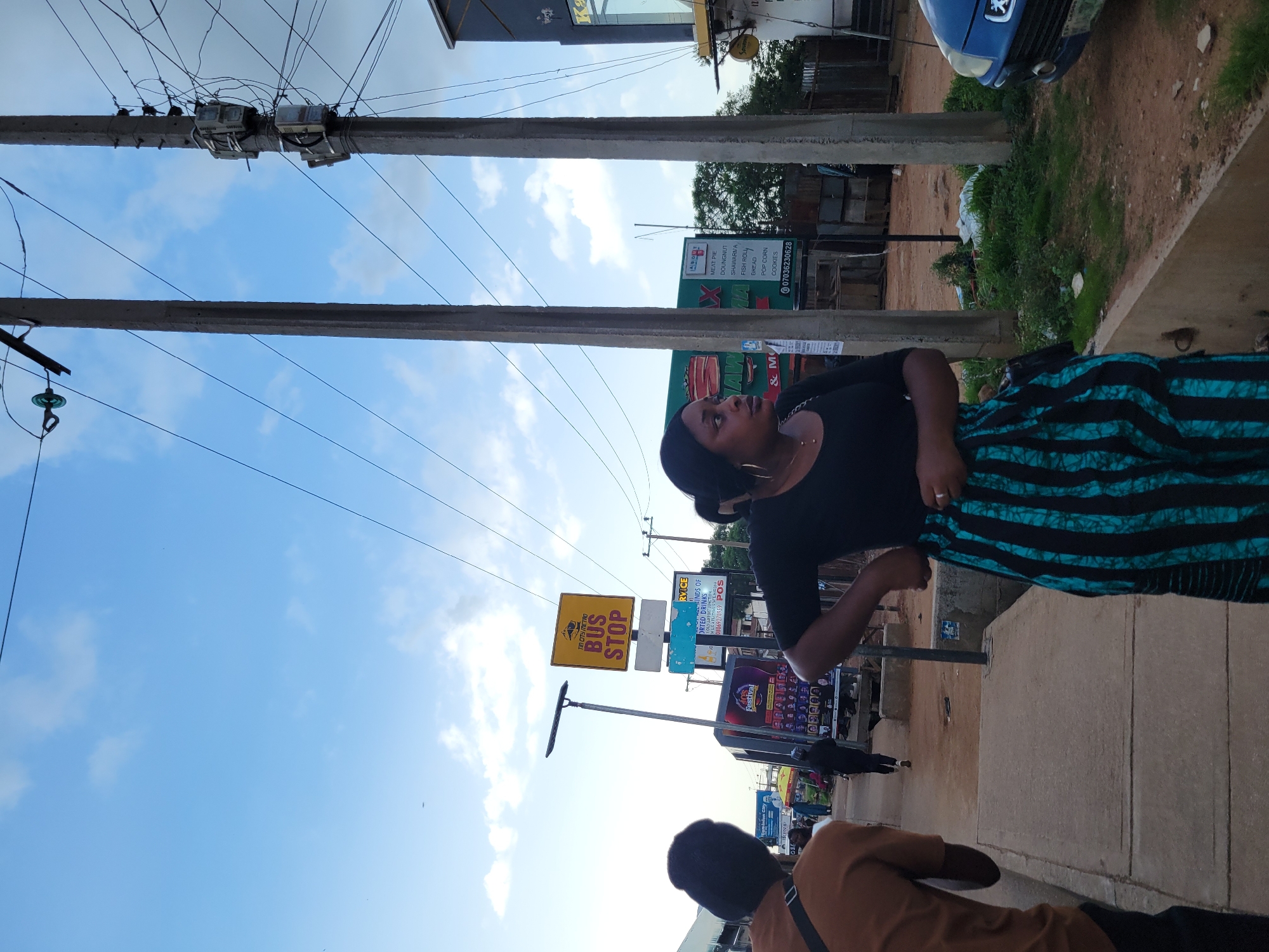 The image shows a street scene in Jos, Nigeria, with a bus stop sign visible in the background. Two women are in the foreground, with one looking to the right and the other with her back to the camera. The woman on the left is wearing a brown shirt and a black beanie, while the woman on the right is wearing a black top and a blue and black striped skirt. There are several utility poles with electrical wires running across the scene, and some signs with text in English, indicating businesses or services. A blue car is parked on the right side of the road. The ground is a mix of concrete pavement and dirt, with some green vegetation. The sky is partly cloudy.