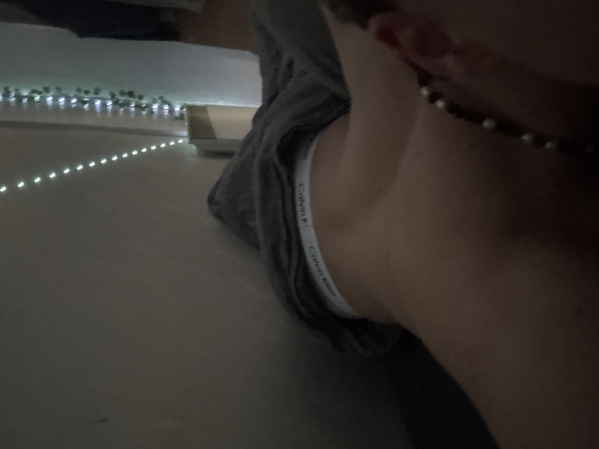 A close-up, low-angle shot shows the neck and shoulder area of a person, likely female. A grey top is visible, revealing the white waistband of Calvin Klein underwear. A necklace with pearls is around the neck. The background is out of focus but shows lights, some in a line, and some in a bunch. The location and specific context of the image within Troyan, Bulgaria, are not discernible.