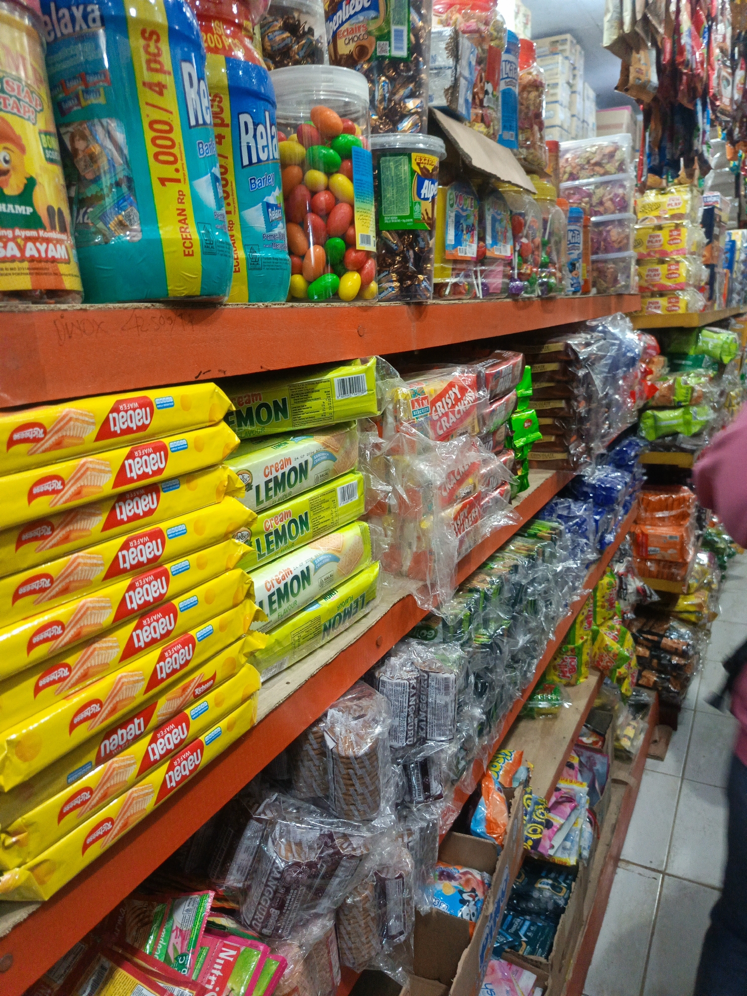 The image depicts a shelf in a small Indonesian convenience store, likely in a neighborhood setting such as Ilotidea. The shelf is stocked with a variety of packaged snacks and drinks, predominantly biscuits, candies, and beverages. Prominently displayed are stacks of yellow packaged "Nabati Wafer" biscuits and green packaged "Relaxa" lemon flavored candies. Behind these, a diverse assortment of other items is visible, including what appear to be chocolate bars, other wafer products, and various individually wrapped snacks. The lighting is somewhat dim, suggesting an indoor environment. No people are clearly visible in the frame, though a blurred figure in a pink top is partially visible on the far right. The shelves themselves are made of wood, with an orange painted metal support structure. The overall impression is of a well-stocked, albeit modestly organized, retail space.