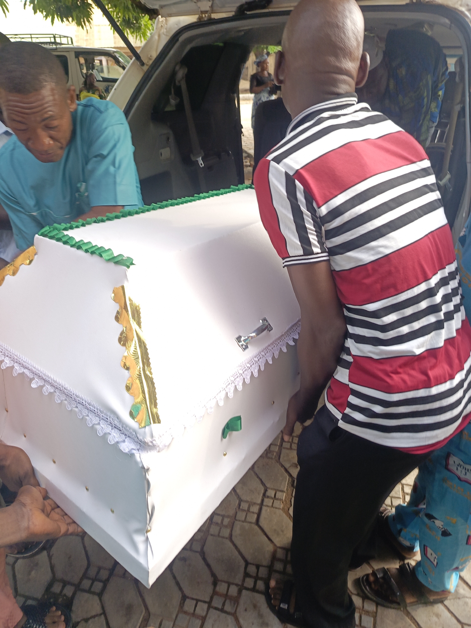 The image captures an outdoor scene in Akwanga, Nigeria, during daylight hours, centering on the handling of a white casket. Several individuals are involved in moving the casket to or from the rear of a white vehicle, likely a van or SUV, which has its hatch open.
In the left foreground, a man in a teal/light blue short-sleeved shirt is visible, looking down as he supports the casket. On the right, a bald man, seen from behind, wears a red, black, and white striped polo shirt, also assisting with the casket. The lower legs of a child in blue patterned pants, with "JOHN..." partially visible on them, are seen on the far right.
The casket itself is the prominent object, covered in white material and elaborately decorated. It features green ruffled ribbon trim along its top edges, gold patterned trim on its sides, and white lace detailing around its base. A silver-colored handle is visible on the casket's lid. The ground is paved with hexagonal tiles. In the background, other people and foliage are faintly visible, along with another vehicle. The scene suggests a funeral or a related ceremonial event.