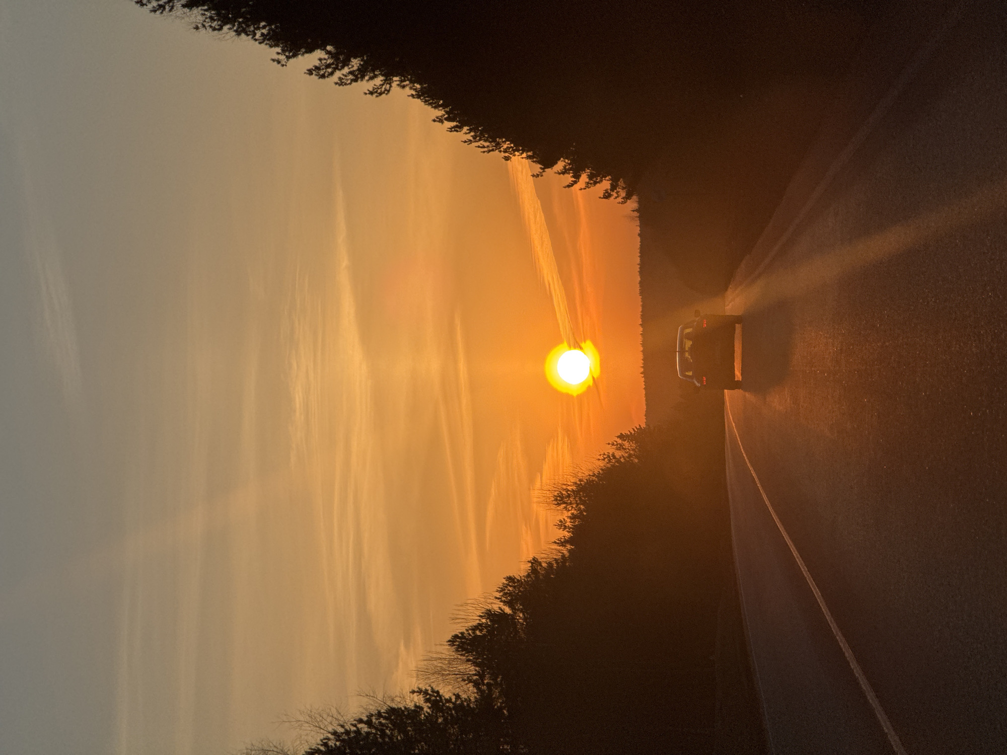 The image shows a car traveling down a road at sunset in Chaikovsky, Russia. The sun is positioned directly ahead, casting a bright glare. The surrounding sky is a mix of orange and yellow hues, and there are trees bordering both sides of the road.