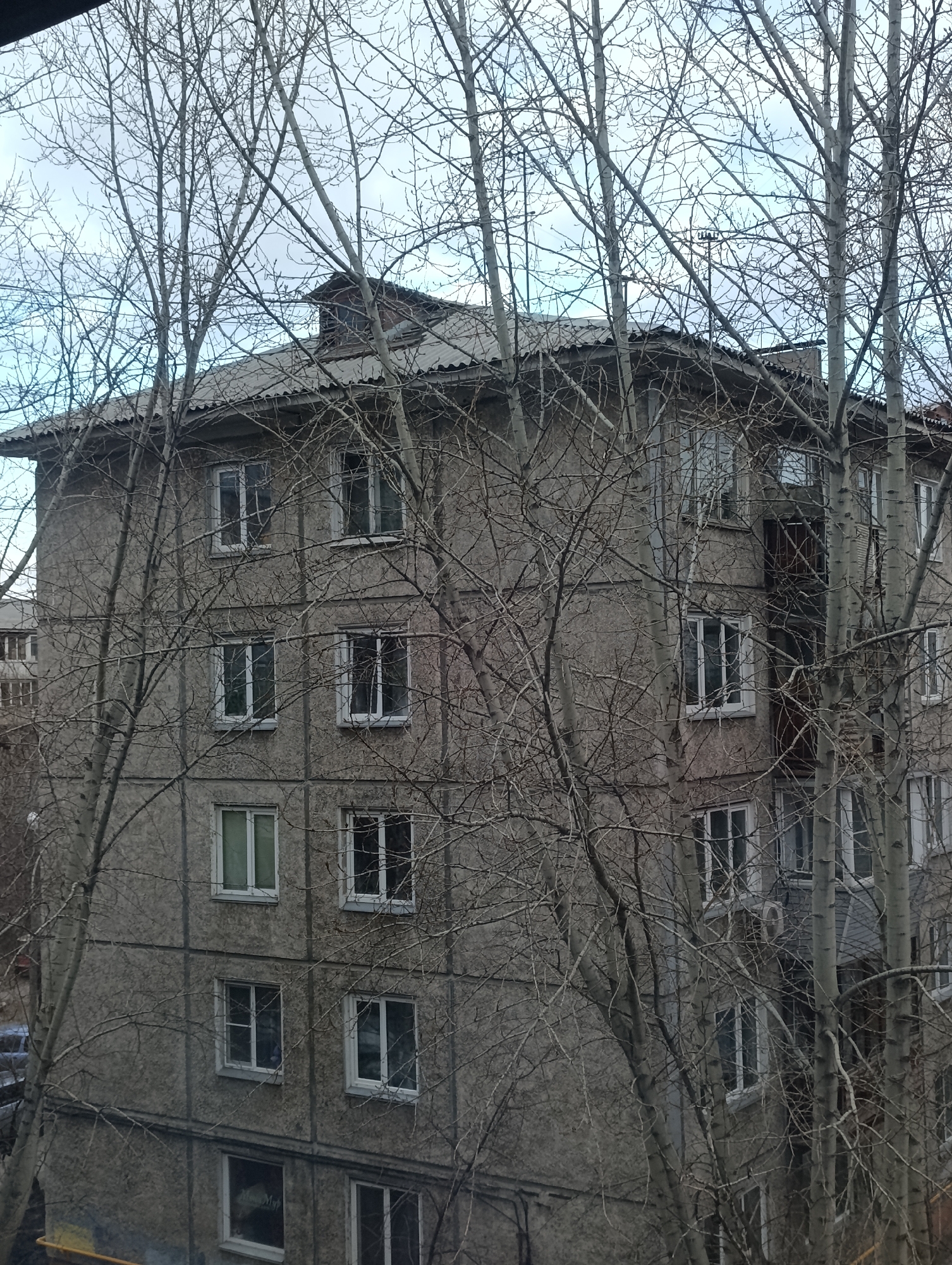 The image shows an old, multi-story residential building in what is likely a residential area of Krasnoyarsk, Russia. The building has a grey concrete exterior with multiple windows. Several bare trees obscure the view of the building. The sky is overcast. No people are visible in the image. A second building is partially visible in the background.
