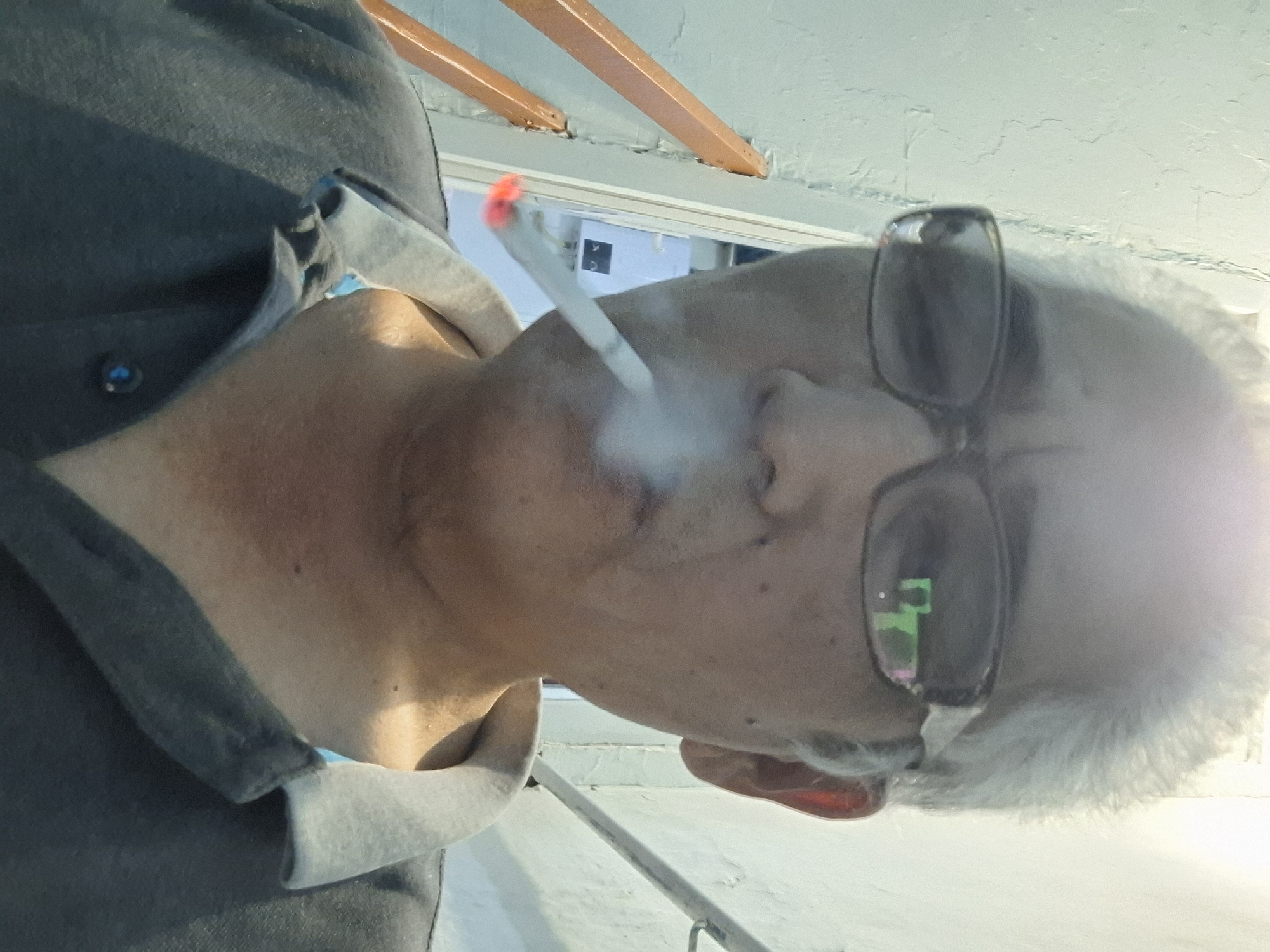 The image is a close-up, head-and-shoulders selfie of an older East Asian man with gray hair, wearing glasses and a dark polo shirt. He is smoking a cigarette that is visible in the lower left quadrant of the frame, with smoke rising from it. The background is out of focus and appears to be an indoor setting with a textured wall. There are no discernible landmarks or elements that would indicate a specific location within the city of Almaty, Kazakhstan.