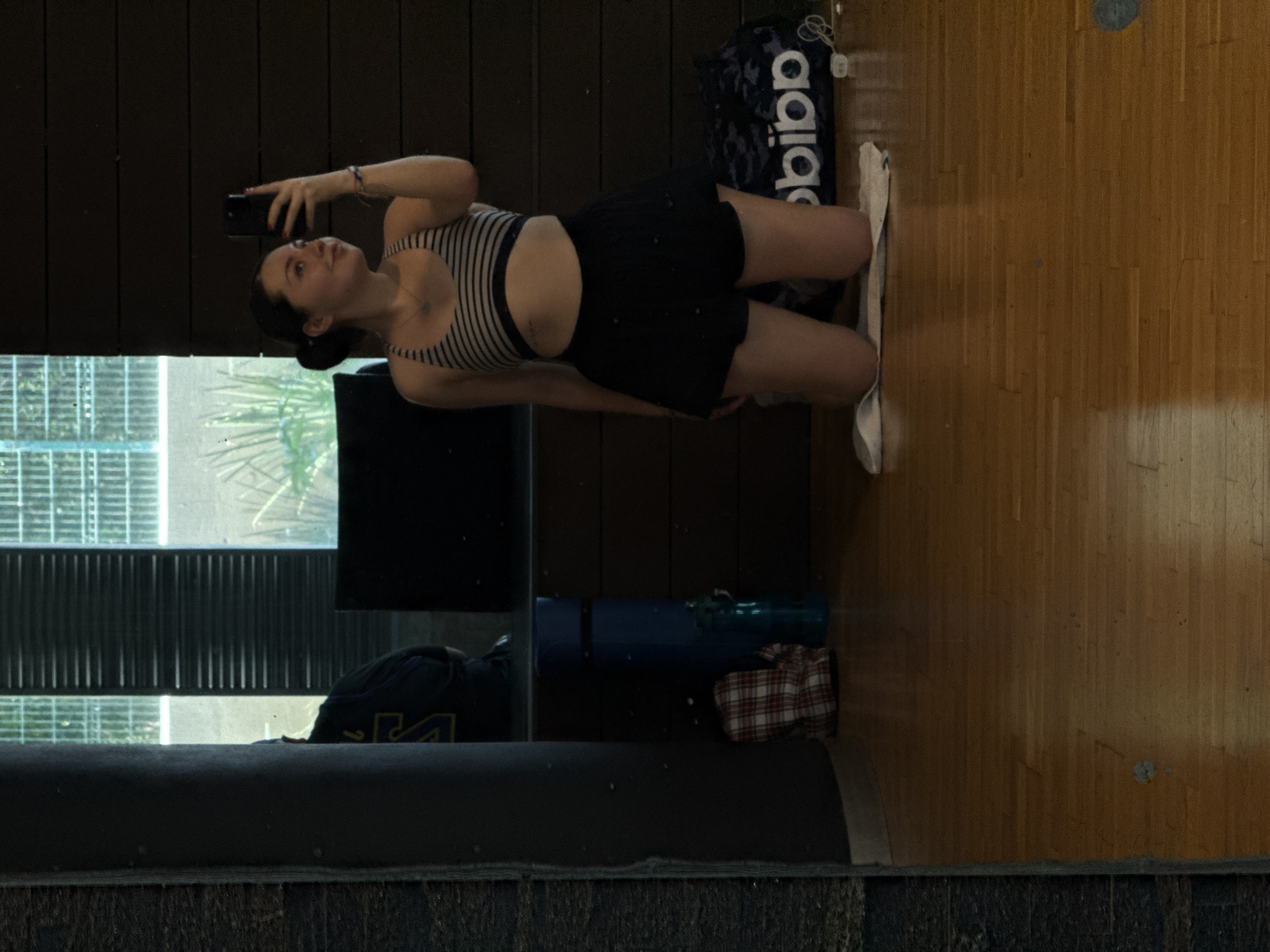A young woman is captured in a mirror selfie, posing in an indoor setting. She is seen standing, slightly bent at the knees, holding a dark-colored smartphone in her right hand, angled to take the photo. Her hair is styled in a bun, and she wears a striped sports bra or crop top, dark shorts, a thin necklace, and a bracelet on her left wrist.
The background reflects a room with dark paneled walls and a wooden floor. To her right, a black Adidas sports bag with "bibb" (part of the Adidas logo) visible in white lettering rests on the floor, next to a white towel or piece of cloth. Further down and to the left, a blue yoga mat and a blue water bottle are placed on the floor, with a red and white plaid shirt beneath them.
The mirror also reflects an outdoor scene through a window in the upper left, showing what appears to be buildings or balconies and some greenery, possibly a palm tree, suggesting an urban environment during daylight hours. In the immediate foreground, a dark, textured surface, possibly a piece of furniture or the edge of the mirror frame, is visible. The woman has a calm expression with a slight smile. The overall impression is a casual, everyday moment, possibly before or after a workout. The location is Mexico City, Mexico.