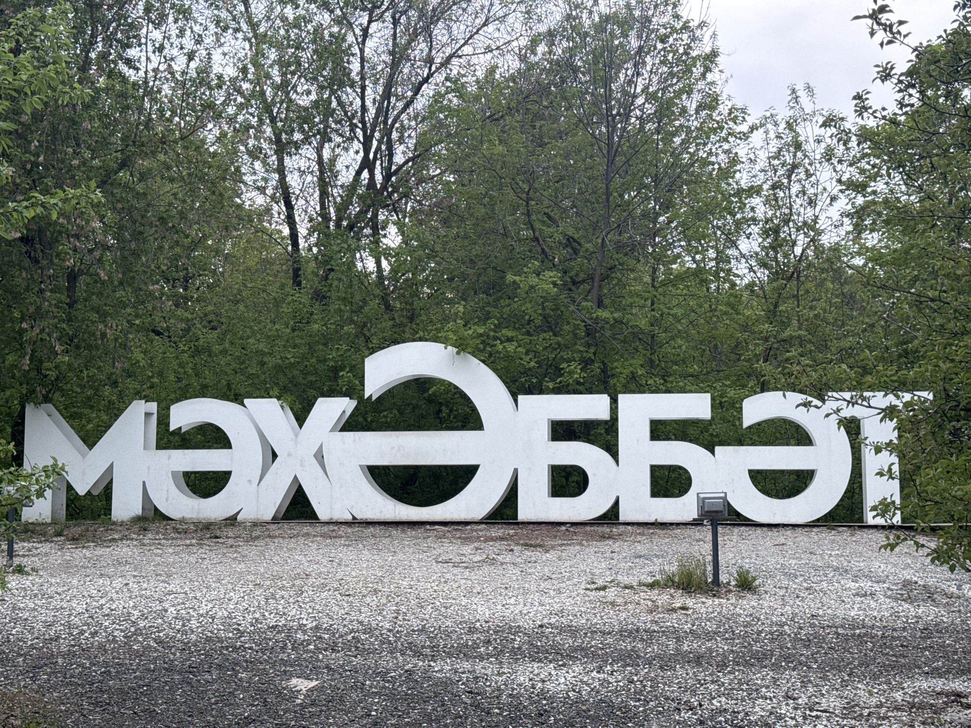A ground-level, medium shot of a large, white, 3D sign displaying the word "МӘХӘББӘТ" (pronounced "mäxäbbät"), which means "love" in Tatar language. The sign is located in Kazan, Russia, as confirmed by the Tatar language. The sign is situated on a gravel surface, with trees and greenery forming a backdrop. The sky is overcast. A small, gray metal box on a pole is visible in front of the sign, slightly to the right of center.