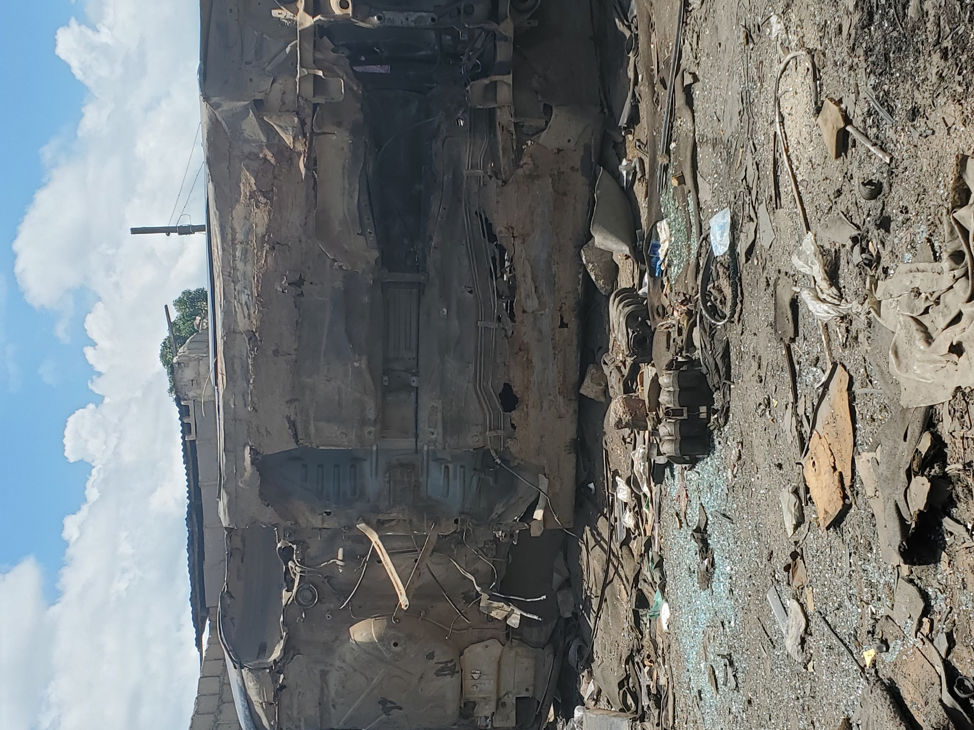 The image depicts a scene of destruction in Rafin Par, Nigeria. A heavily damaged vehicle, appears to be a truck or bus, lies on its side, its metal chassis and interior exposed. The vehicle is severely mangled, with large sections torn open, revealing internal mechanics and structural components. Wires and debris are scattered around the vehicle.
The ground is covered in rubble, including broken glass, dirt, and various pieces of debris. Some fragmented objects that could be parts of other vehicles or structures are visible. In the background, a brick wall and a corrugated metal roof suggest the presence of buildings or structures that may have also been affected by the incident. The sky is partly cloudy with white clouds against a blue sky. The overall scene conveys a sense of recent destruction and chaos.