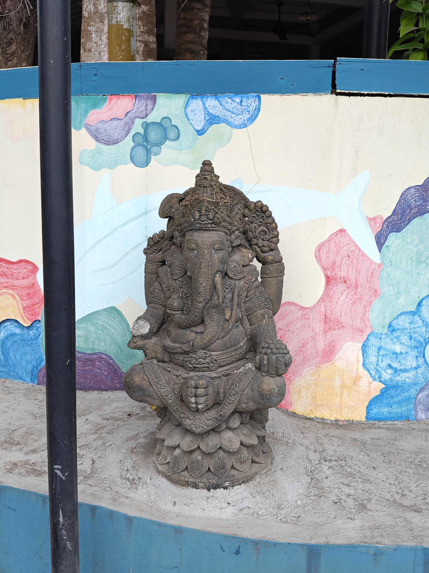 A stone statue of Ganesha sits on a concrete base in front of a painted wall. The wall features various colorful designs, including a large white dolphin. A black pole stands to the left of the statue. The scene is located in Nusa Penida, Indonesia.