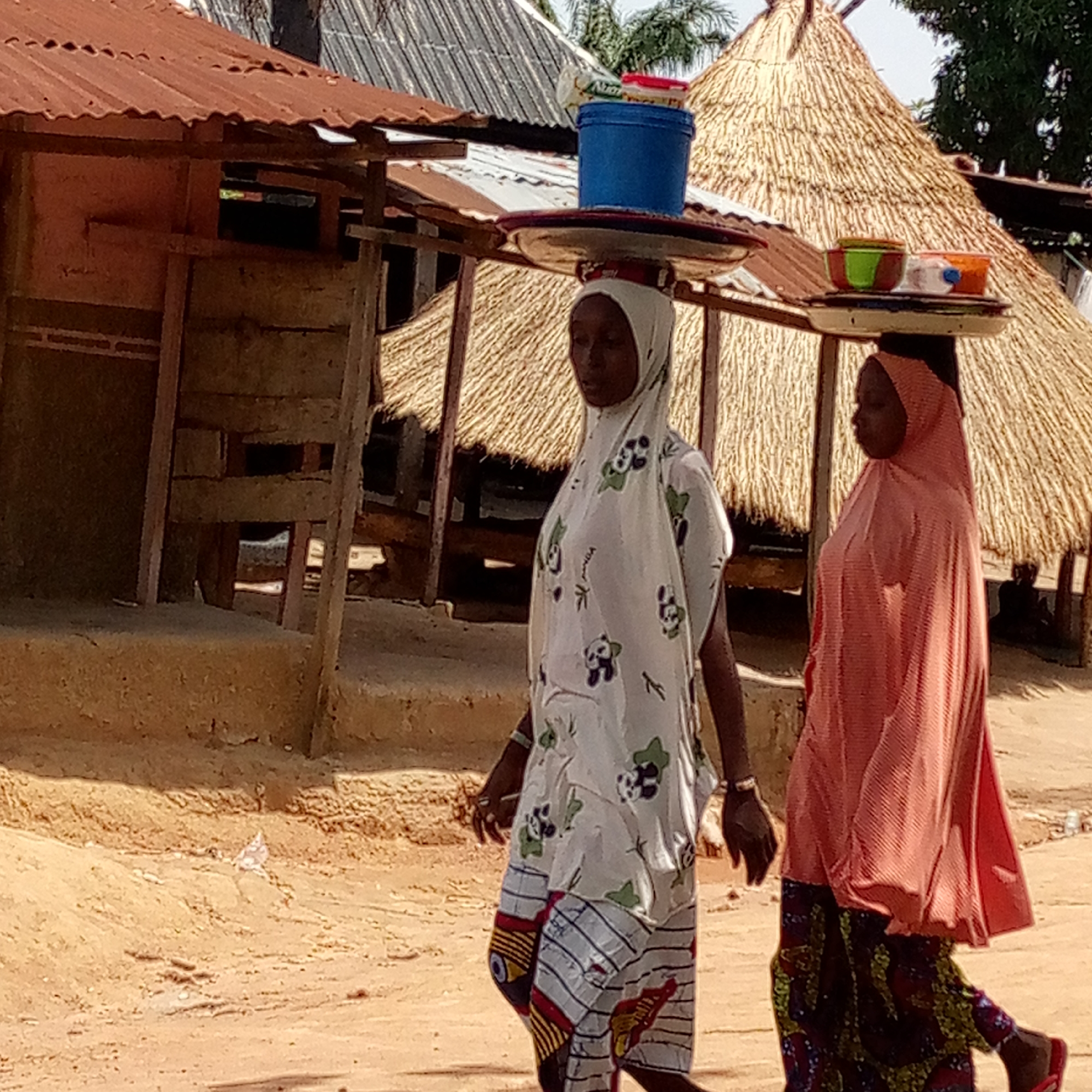 Two women are walking on a dirt road in Mararaba, Nigeria. Both women are carrying items on their heads. One woman, wearing a white patterned headscarf and dress, is carrying a tray with a blue bucket on top. The second woman, wearing a peach-colored headscarf and dress, is carrying a tray with bowls on top. In the background are huts with thatched roofs and metal roofs.