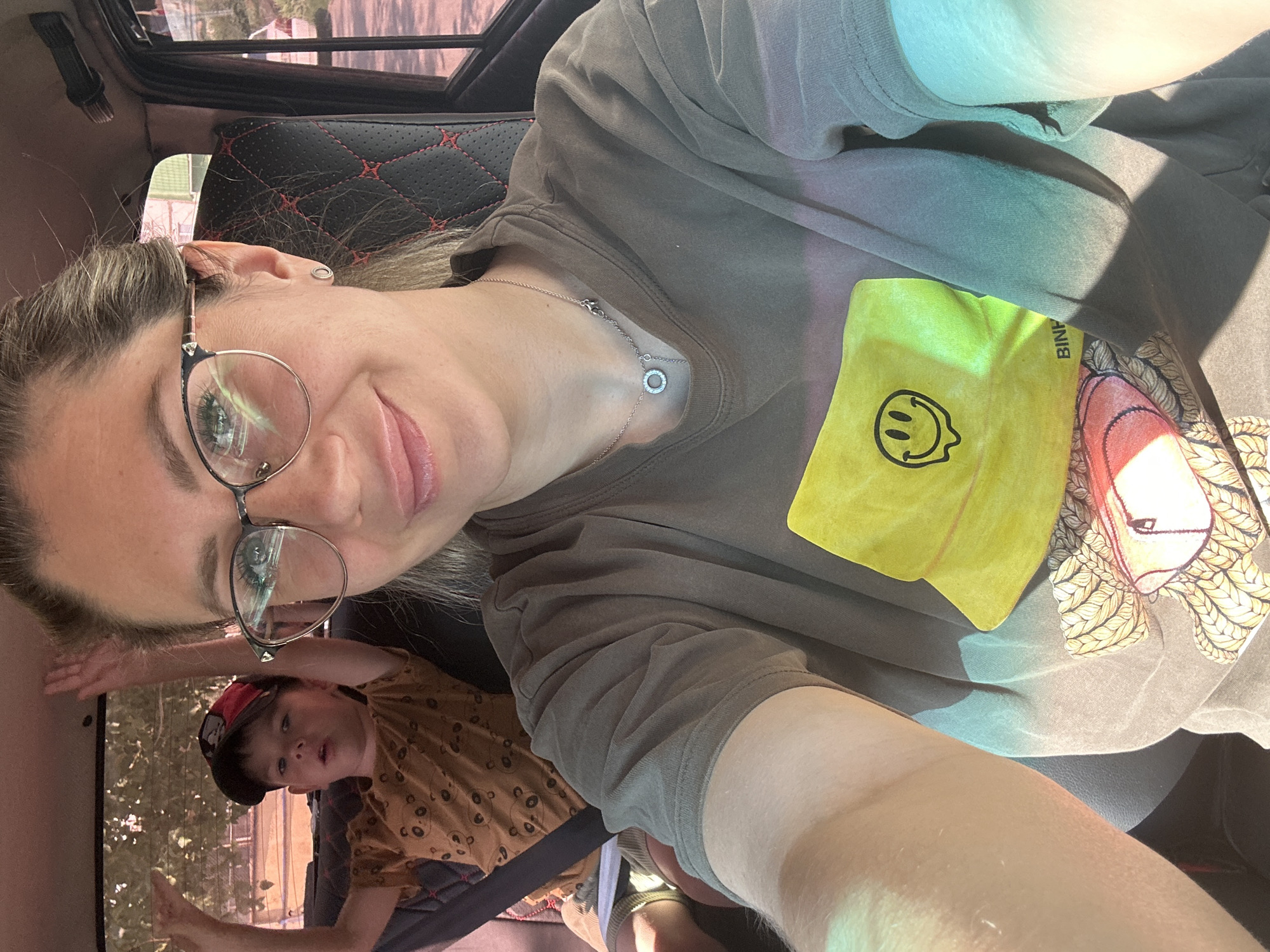 In the back seat of a vehicle, a woman in glasses smiles at the camera while a young boy in a cap and patterned shirt sits behind her, with his hands raised. The scene appears to be during the day. The location is Novoaltaysk, Russia.