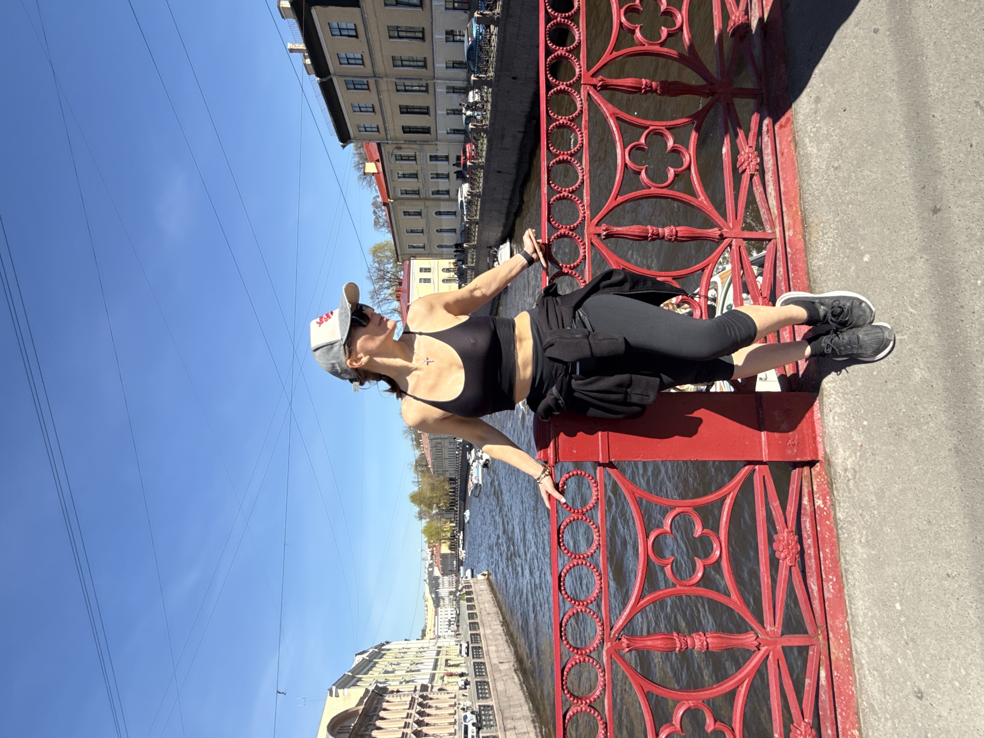 A woman is lying on a red bridge in Saint Petersburg, Russia. She is wearing a black top and black leggings with a grey baseball cap and black sneakers. She is facing the water and resting her hands on the railing. The scene also includes a body of water with boats, buildings in the background, power lines, and a clear blue sky.
