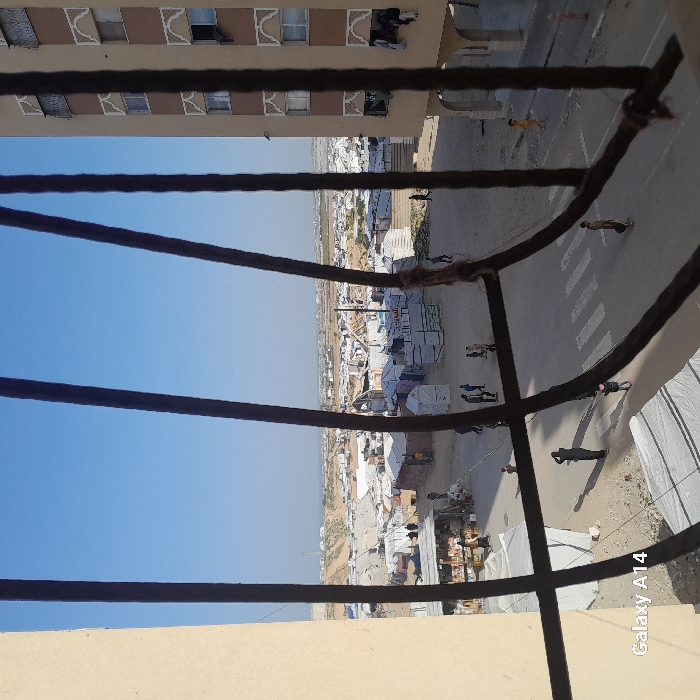 The view from a high window reveals a landscape of tents and temporary structures, possibly a refugee camp or settlement, stretching into the distance under a clear sky. A road with pedestrian crossings cuts through the scene, with figures of people walking along it. The foreground features a metal window grill, while a building with multiple windows occupies the top portion of the frame.