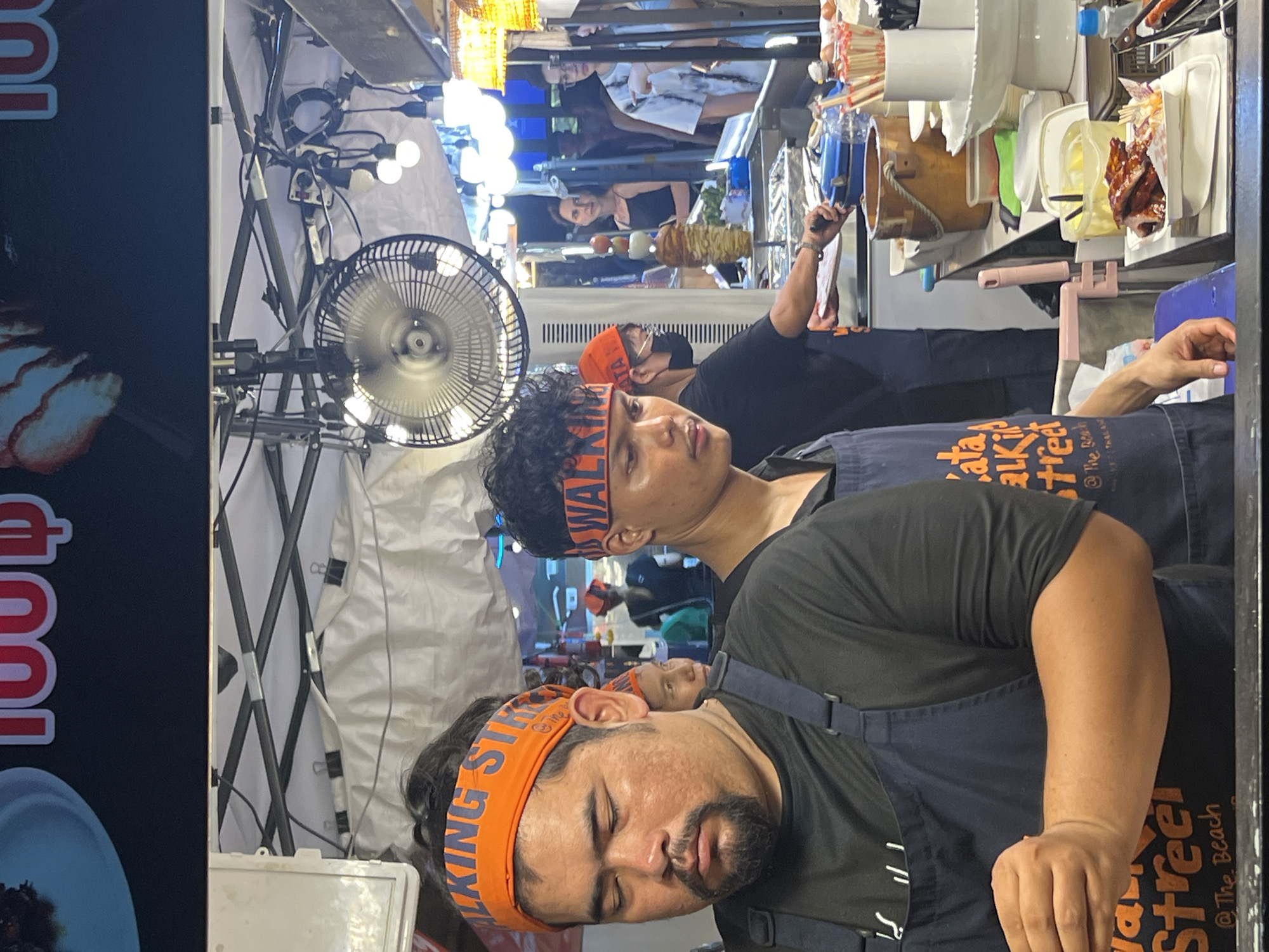This image depicts a bustling street food stall in Karon, Thailand, likely during the evening given the artificial lighting. The scene is filled with people and food preparation items. In the foreground, two individuals are visible. One man, wearing an orange headband with the text "WALKING STREET," is lying down with his eyes closed, appearing to be asleep or resting. Another person, also wearing a similar orange headband and an apron, is partially visible behind him. The background shows the operational side of the food stall, with cooking equipment, containers of ingredients, and various utensils. A small electric fan is mounted to cool the area. The overall atmosphere seems lively, characteristic of a night market or street food scene. Visible text includes "WALKING STREET," "KATA," "TALKIN STREET," and "@The Beach," suggesting the stall is part of a popular tourist area. The food being prepared is not entirely clear, but what appears to be grilled meat is visible in a container.