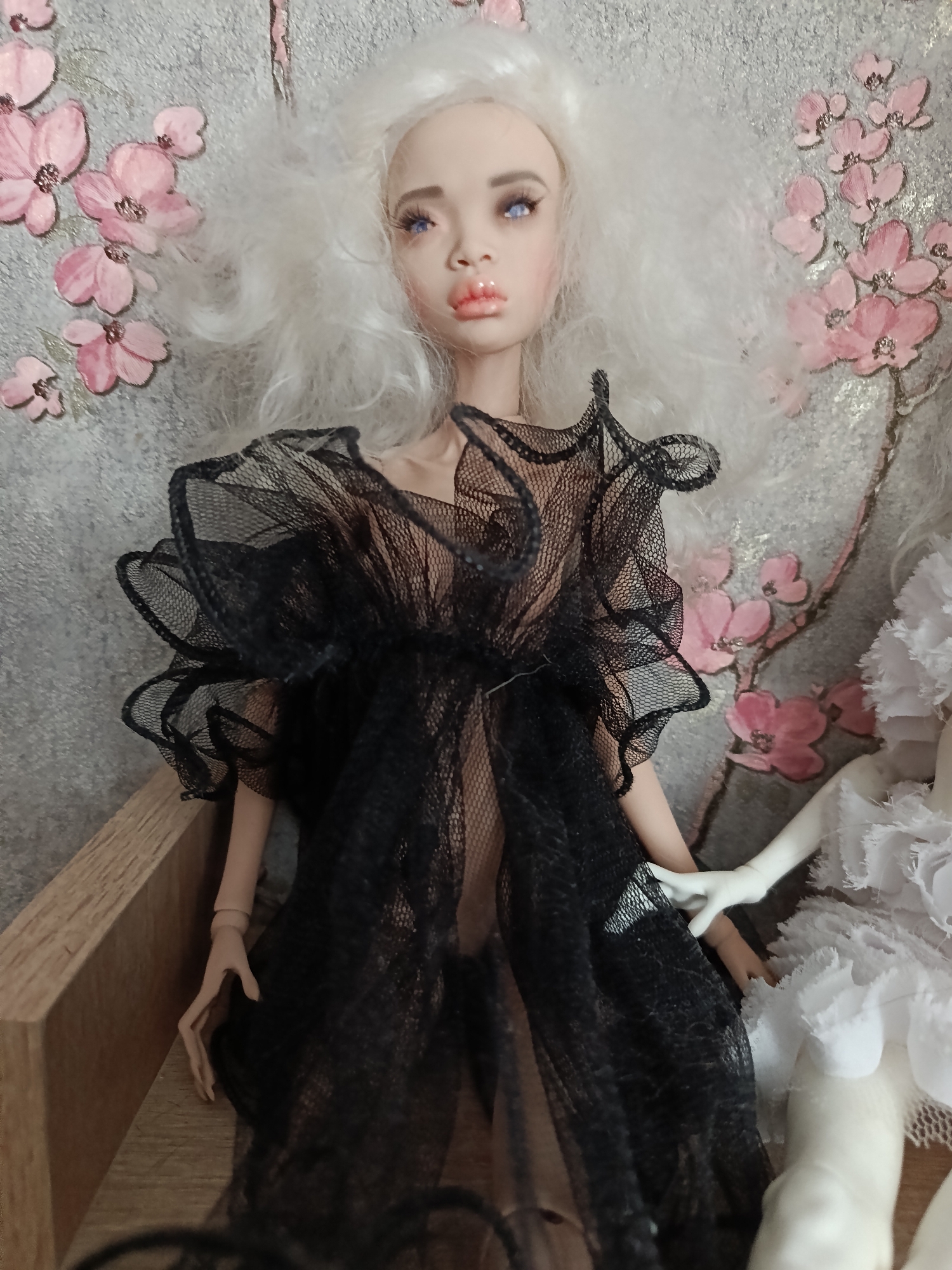 This media file shows a close-up of a doll with white, wavy hair and blue eyes. The doll is wearing a sheer black dress with ruffled sleeves. The background features a textured gray surface adorned with painted pink cherry blossoms. The doll is positioned on a wooden surface, with a partial view of another doll in a white dress to its right. The overall aesthetic is delicate and artistic, possibly representing a theatrical or fantasy scene. The setting appears to be indoors, with soft lighting that highlights the doll's features and the details of its attire and surroundings. There is no visible text in the image. The location context provided is Saint Petersburg, Russia, but this cannot be visually confirmed from the image itself.