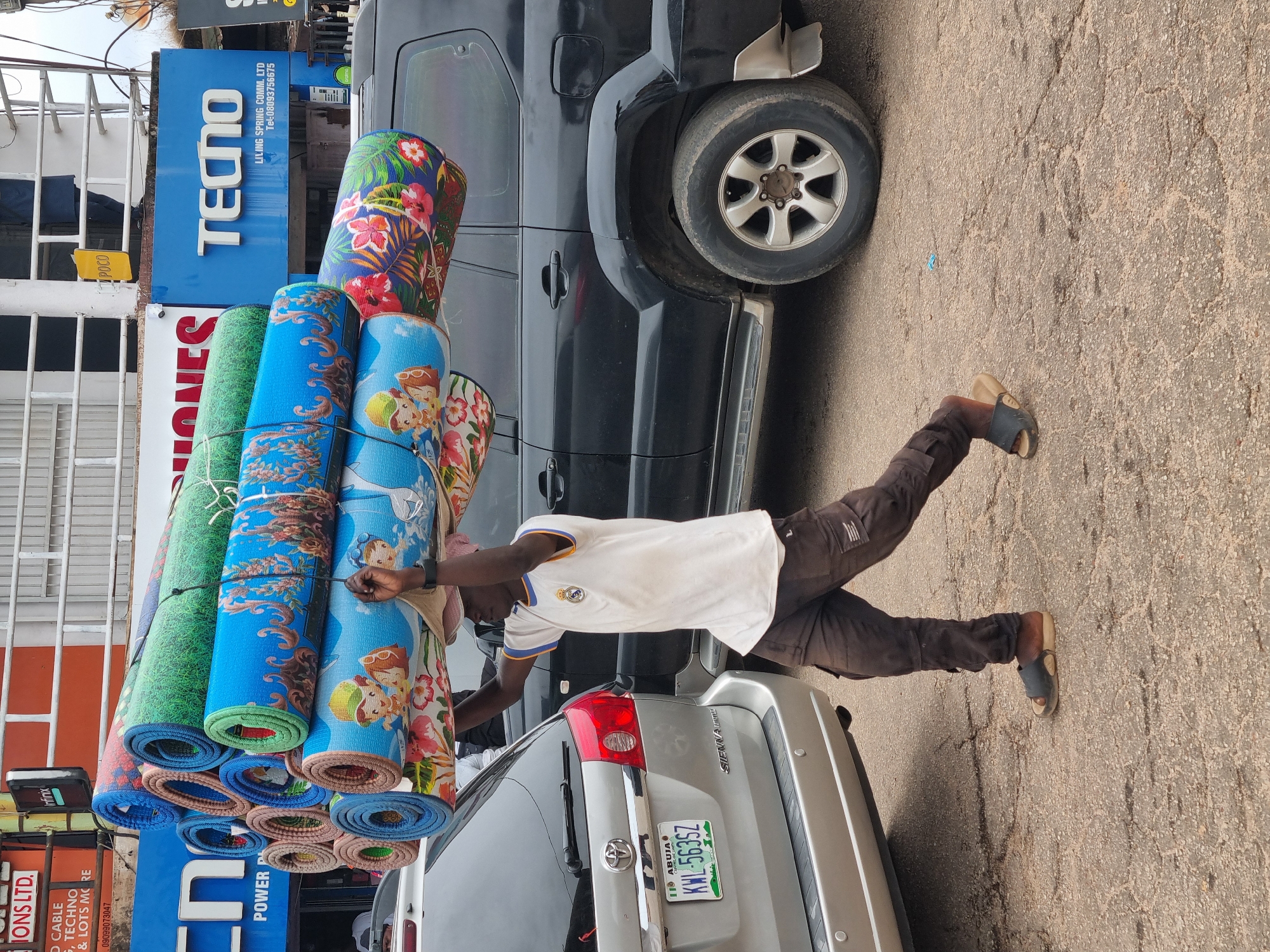 The image shows a man carrying a stack of rolled-up mats on his head and shoulders next to a silver Toyota Sienna SUV with a Nigerian license plate (KWL-563SZ) that reads “Abuja”, near a black SUV. The man wears brown pants, a white shirt, and sandals. Behind the vehicles are storefronts with signage. One sign reads "TECNO, LIVING SPRING COMM. LTD." and includes a phone number. Another sign reads "IONS LTD. CABLE, TECHNO." The location appears to be in Nigeria, likely based on the license plate on the silver vehicle. The location is not Jos, Nigeria, as indicated in the instructions, but rather Abuja, Nigeria, based on the license plate on the car. The street is paved and appears to be in an urban area.