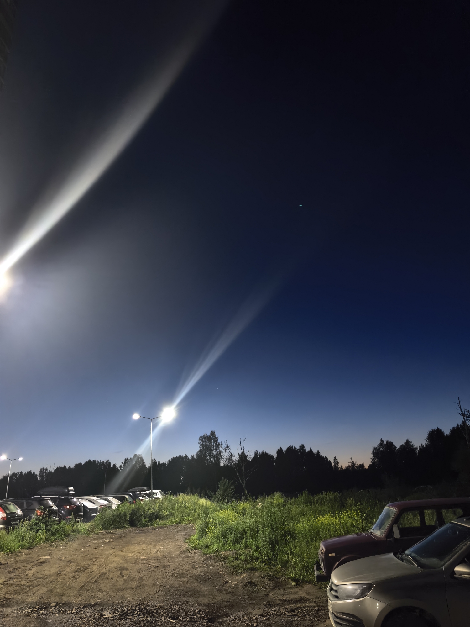 The image depicts a nighttime scene in an outdoor area, possibly a parking lot or a rural roadside, located in an unspecified region. A clear, deep blue sky dominates the upper portion of the frame, with a gradient towards a lighter blue near the horizon. Two bright, powerful light beams cut across the sky from left to right, suggesting the presence of streetlights or floodlights. Faint, wispy clouds or atmospheric effects are visible within these light beams, adding texture to the night sky.
Below the sky, a dense line of dark trees forms a silhouette against the horizon, indicating a wooded area. In the foreground and middle ground, a dirt and gravel surface suggests a path or unpaved area. To the left, a row of parked cars is visible, some with roof racks, lined up along the edge of the wooded area. To the right, two cars are parked closer to the viewer, one a dark red older model vehicle and the other a lighter colored modern sedan. The surrounding vegetation is lush and appears to be tall grass and wildflowers, illuminated by the artificial light. The overall impression is one of a dimly lit, open space on the outskirts of civilization.