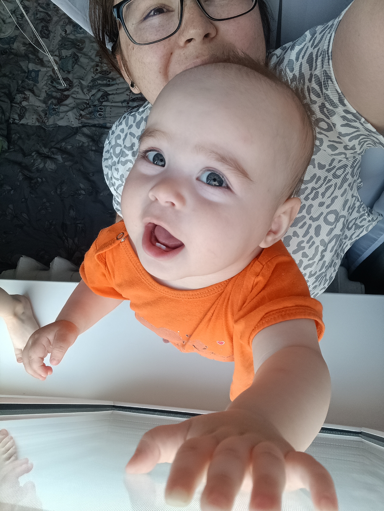 A close-up, top-down shot shows a baby reaching towards the camera. The baby is wearing an orange onesie and is positioned in the foreground. Behind the baby, a woman wearing glasses and a patterned shirt is partially visible. The background consists of a patterned blanket and what appears to be a white surface. The lighting suggests an indoor environment. There is no identifiable information about the specific location in Noviy, Russia.