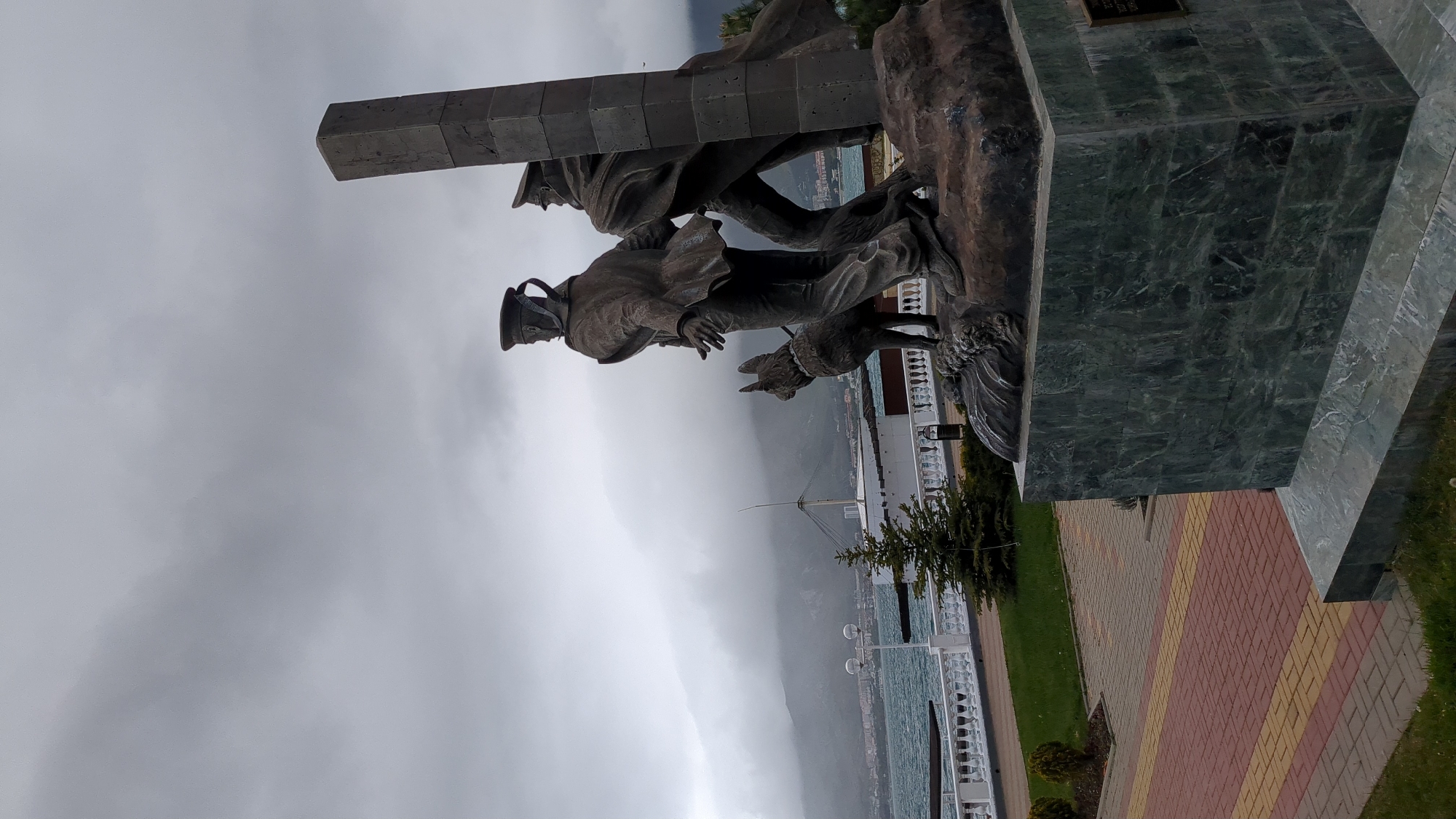 The image shows a bronze monument of two soldiers and a dog, located in Gelendzhik, Russia. One soldier carries a beam on his shoulder while the other, wearing a cap, leads a dog by his side. The monument stands on a large marble base on a paved walkway near the coast. In the background are the sea and a low mountain range under a cloudy sky.