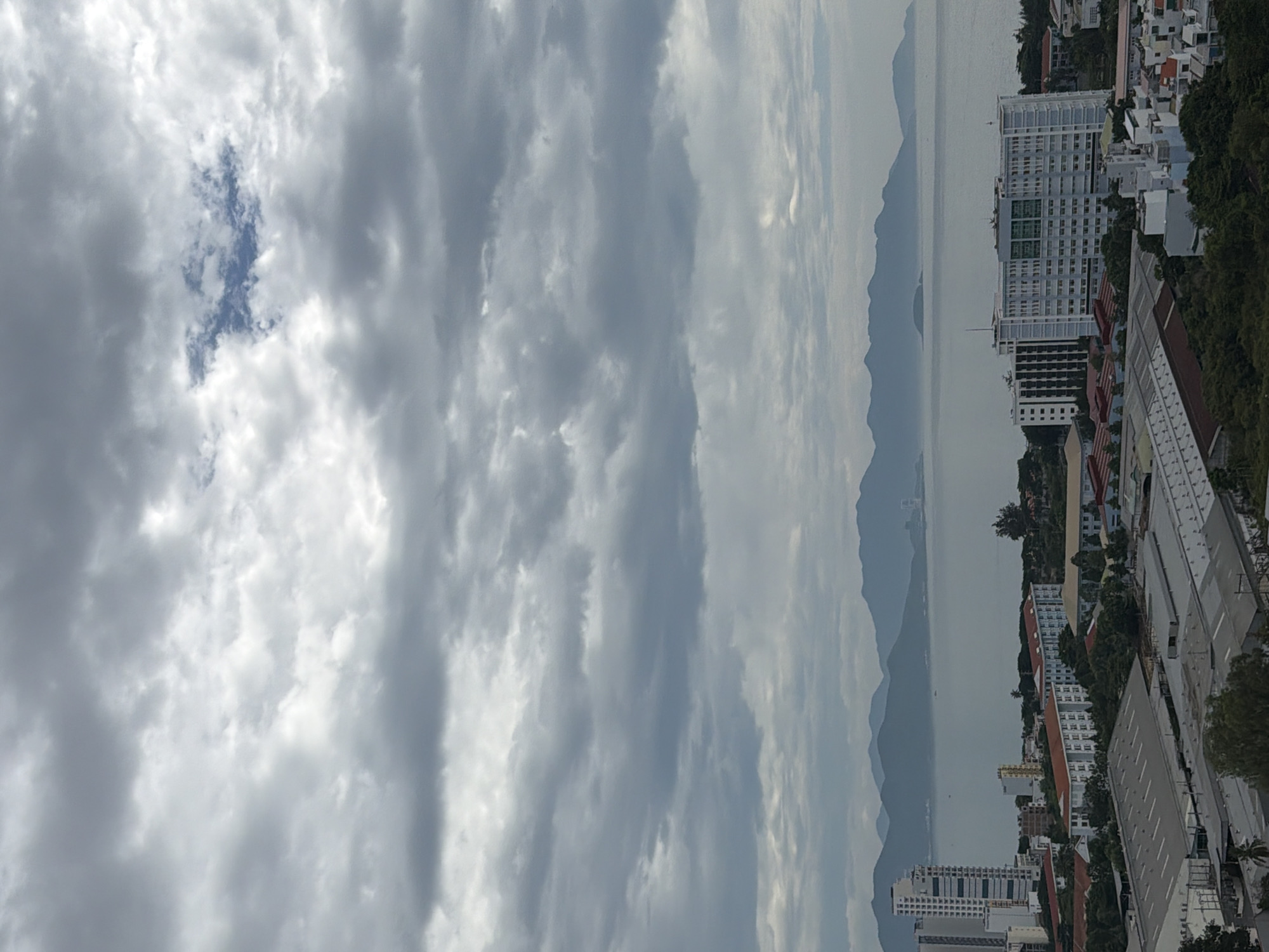 The image captures a wide shot of the city of Nha Trang, Vietnam, under a cloudy sky. In the foreground, various industrial and residential buildings are visible, characterized by their flat roofs and a mix of light and dark colors. Further back, a body of water, likely the sea, separates the city from a series of undulating, mist-shrouded mountains in the distance. A prominent, tall, white building with many windows stands out among the urban landscape. The overall scene suggests a coastal city setting with a blend of commercial and residential development.
