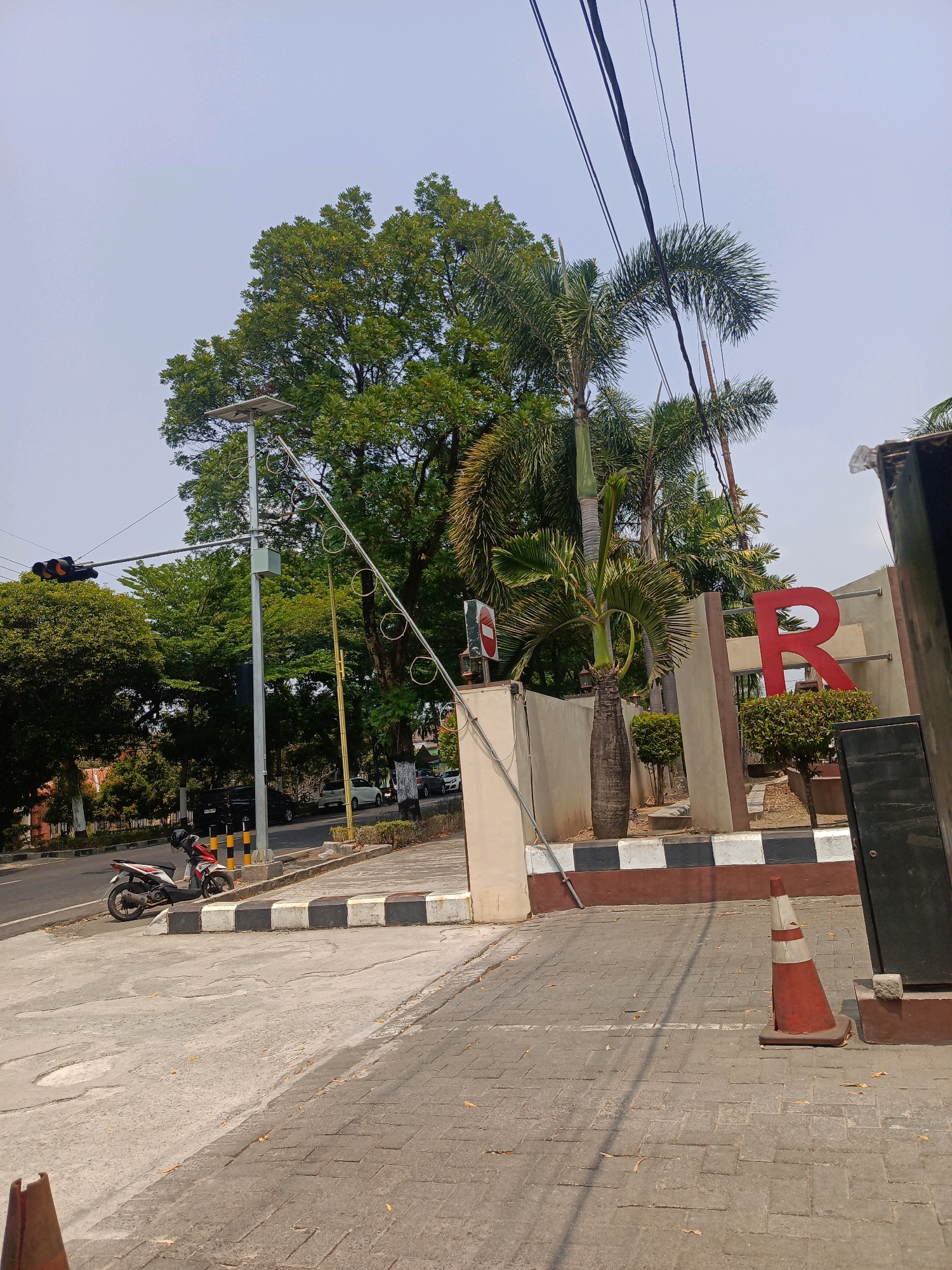 The image depicts a street scene in the city of Tambran, Indonesia. In the foreground, a grey, paved area transitions to a grey sidewalk with a black and white striped curb. A red and white traffic cone stands on the sidewalk. To the left, a white motorcycle is parked on a concrete surface. In the background, a street with several cars is visible. A traffic light with orange lights illuminated is positioned over the road. A tall, silver lamppost with a solar panel on top stands on the sidewalk. Behind the lamppost and extending across the middle ground are several large trees with green foliage, including what appears to be a palm tree. To the right, a building wall with a large red letter "R" is visible, along with some decorative shrubbery. Overhead, a network of black electrical wires crisscrosses the sky. The sky is clear and pale blue.