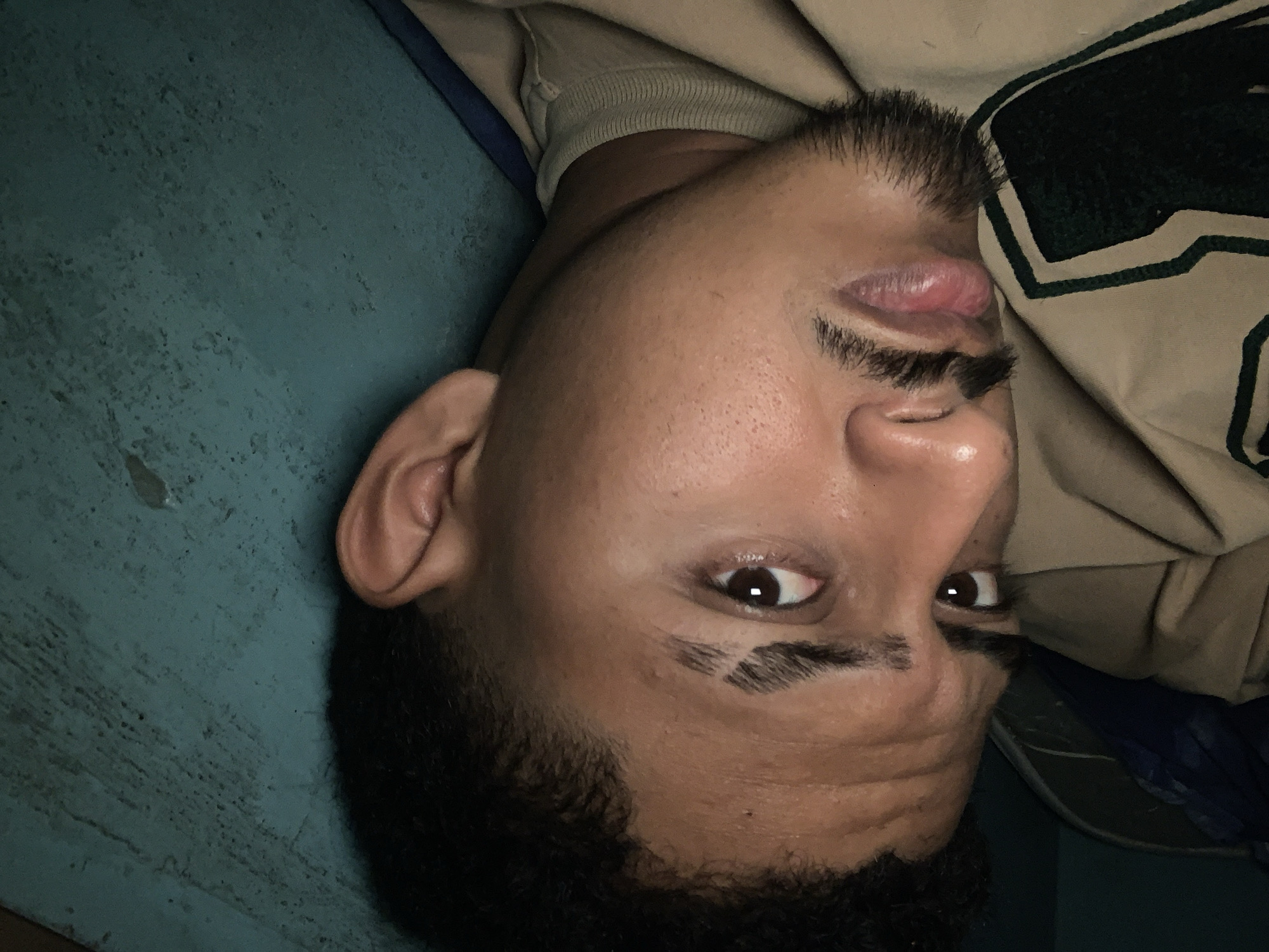 This image is a close-up portrait of a young man, rotated 90 degrees counter-clockwise. He has medium skin tone, dark, curly hair, and dark brown eyes. One eye is wide open and looks directly at the viewer, while the other is partially visible. He has a dark, neatly trimmed mustache and goatee beard. His lips are slightly parted.
He is wearing a light beige or tan long-sleeved shirt with a ribbed cuff visible near his left ear. A portion of the shirt on his right side features a dark green fabric with a white, possibly striped or block pattern.
The background consists primarily of a textured, matte teal or greenish-blue surface, which appears worn or painted, possibly a wall or a mat. A dark blue or black fabric is also visible behind his right shoulder.
The lighting is dim, suggesting an indoor setting or low-light conditions. The man is posing for the camera, gazing directly forward. There is no visible text in the image. While the location context is Santa Ana, Venezuela, no specific geographical cues are visible within the photograph itself.