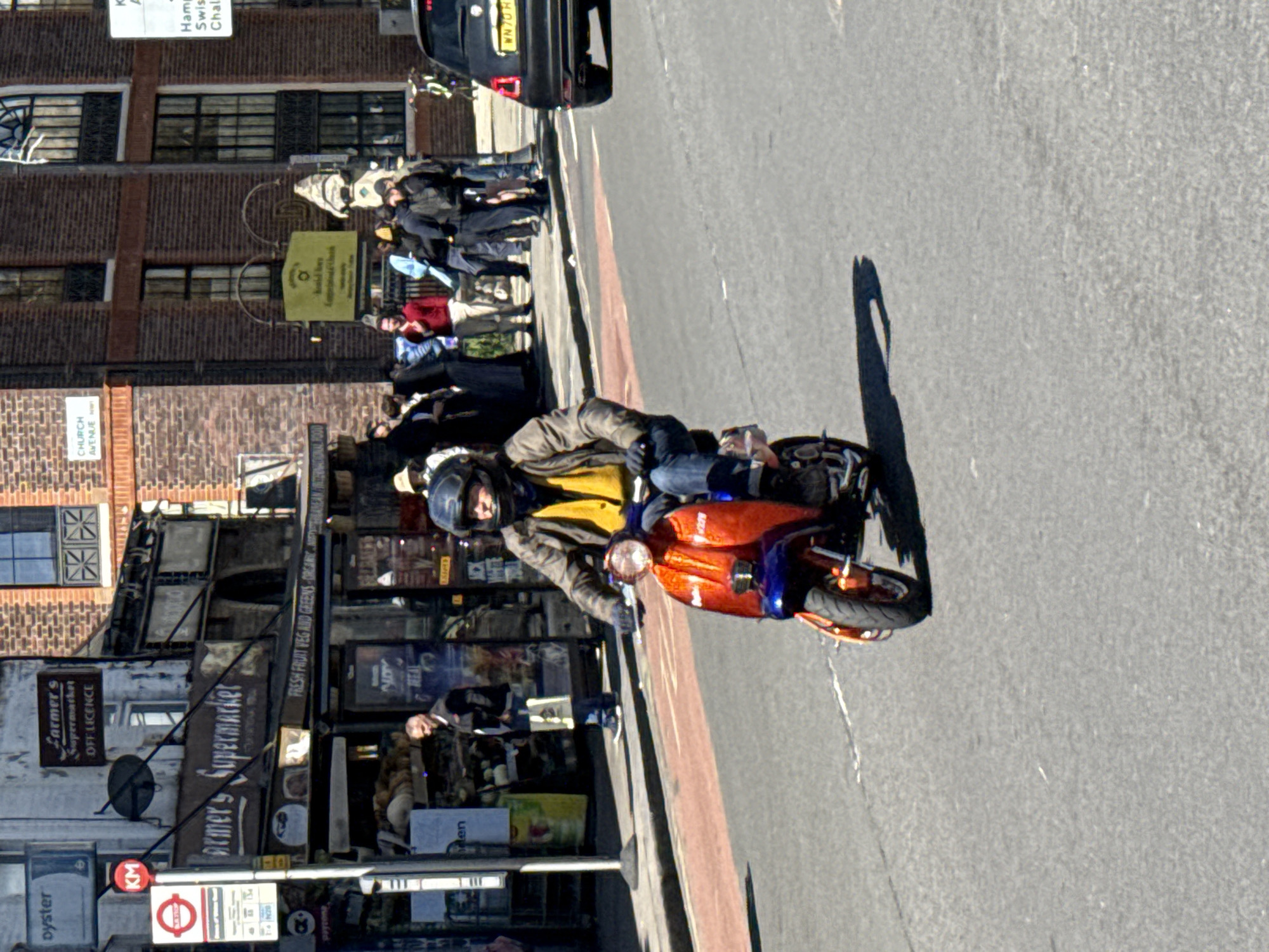 A man wearing a helmet and jacket rides a red and blue moped on a street in London. The street has signs that read "Farmer's Supermarket," "Fresh Fruit Veg and Greens Organic," and "Church Avenue." A bus stop and streetlights are on the left. In the background, a group of people walk on the sidewalk, and a black vehicle is parked at the end of the street. The license plate of the black vehicle reads "WN70 H."