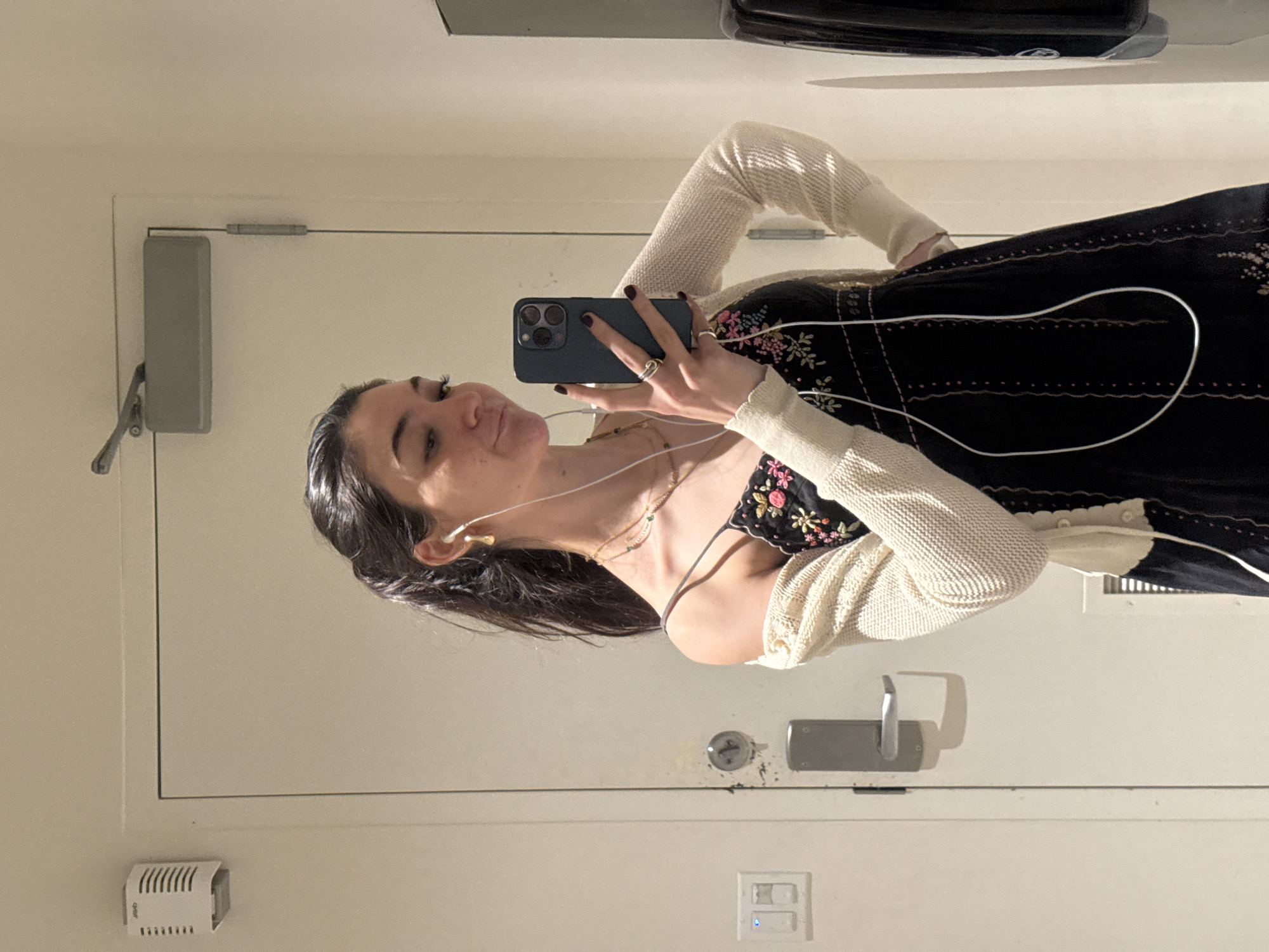 A young woman with dark hair pulled back in a ponytail is taking a mirror selfie in what appears to be a public restroom. She is wearing a black slip dress with embroidered floral details and a cream-colored cardigan. She has white earbuds in and is holding a dark-colored smartphone to take the picture. She is wearing rings on her fingers. The background consists of a closed door with a door handle and a light switch. There is also a white vent on the wall above the door. The overall lighting suggests an indoor setting. The location is not specified to be New York City, United States of America.