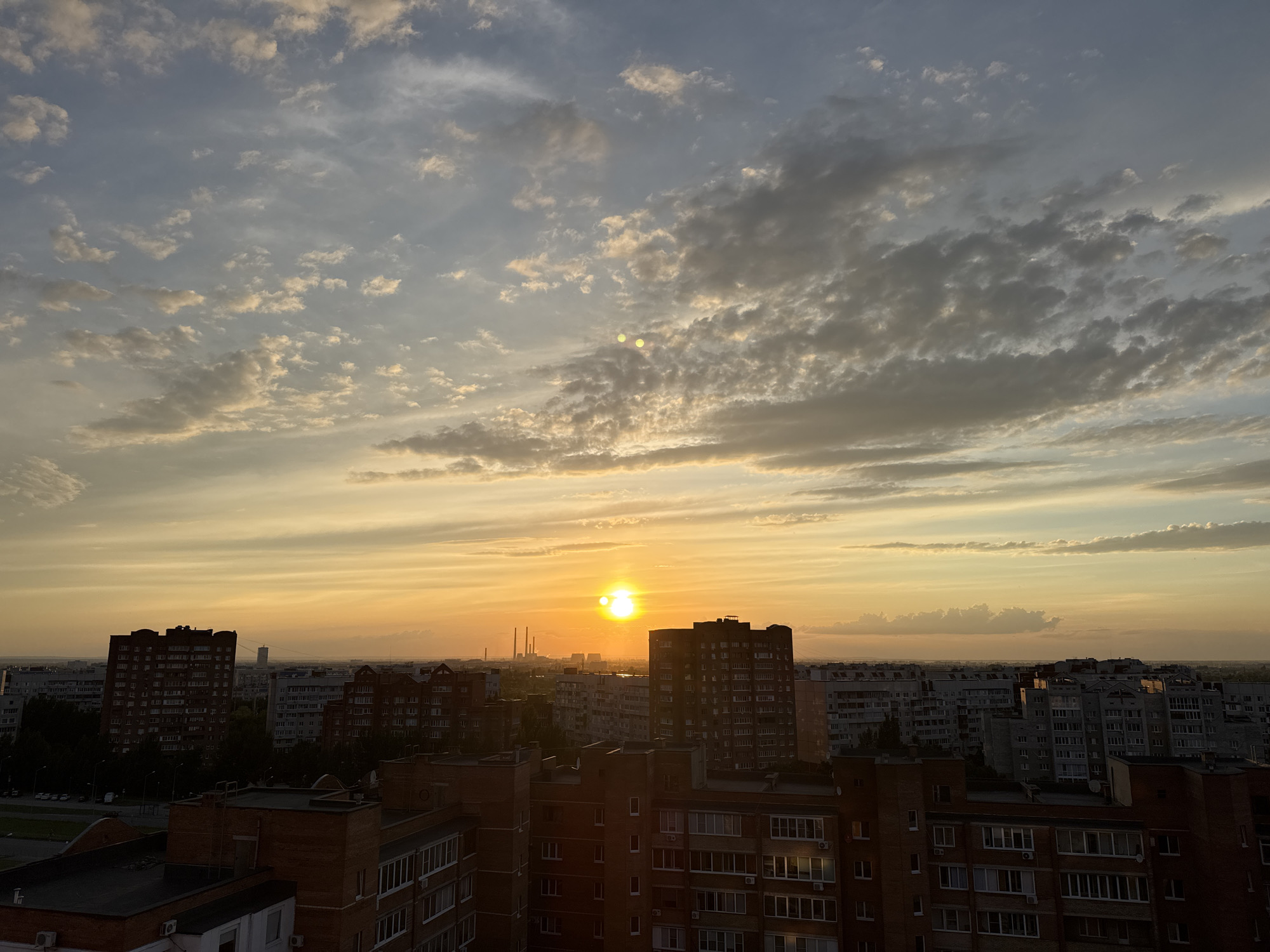 The image depicts a cityscape at sunset. The sun, a bright orb, is positioned slightly left of center, casting a warm, golden glow across the sky. The sky is filled with a mix of light and dark clouds, some tinged with orange and yellow from the setting sun. In the foreground, a dense collection of multi-story buildings dominates the view. These buildings are predominantly residential, with visible windows and balconies. Some structures are constructed from red brick, while others appear to be made of concrete or have lighter-colored facades. In the distance, beyond the immediate buildings, more urban sprawl is visible, with a faint outline of industrial structures, possibly chimneys, visible on the horizon. The overall impression is one of an urban environment bathed in the soft light of dusk.