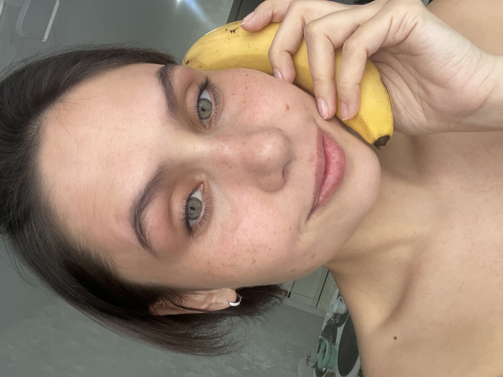 A young woman with fair skin, freckles, and dark brown hair holds a yellow banana to her right cheek. She has green eyes and a small silver earring in her left ear. The background is indistinct, but appears to be an interior space. There is no indication of Izhevsk, Russia.