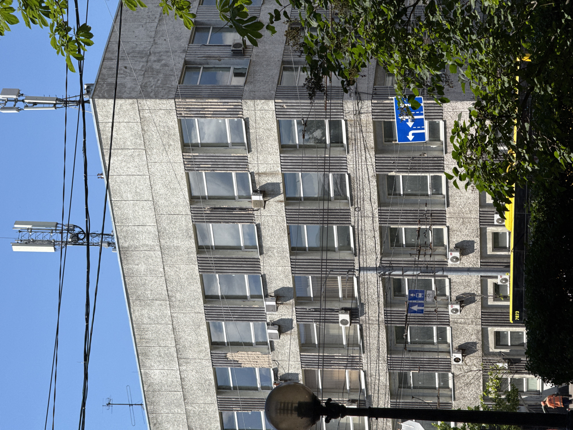 The image shows a modern multi-story concrete building in Kyiv, Ukraine. The building's facade is textured and features rows of windows with dark vertical trim. Several air conditioning units are mounted on the exterior walls. On the rooftop, there are communication antennas. Power lines are strung across the upper portion of the frame, and a street lamp is visible on the left. In the foreground, lush green trees obscure parts of the lower floors. A yellow bus with the number 7873 is partially visible at the bottom. A person in an orange uniform walks on the sidewalk on the left. Two blue traffic signs are mounted on the building, one indicating lane directions and another with a bicycle symbol. The sky is clear and blue.