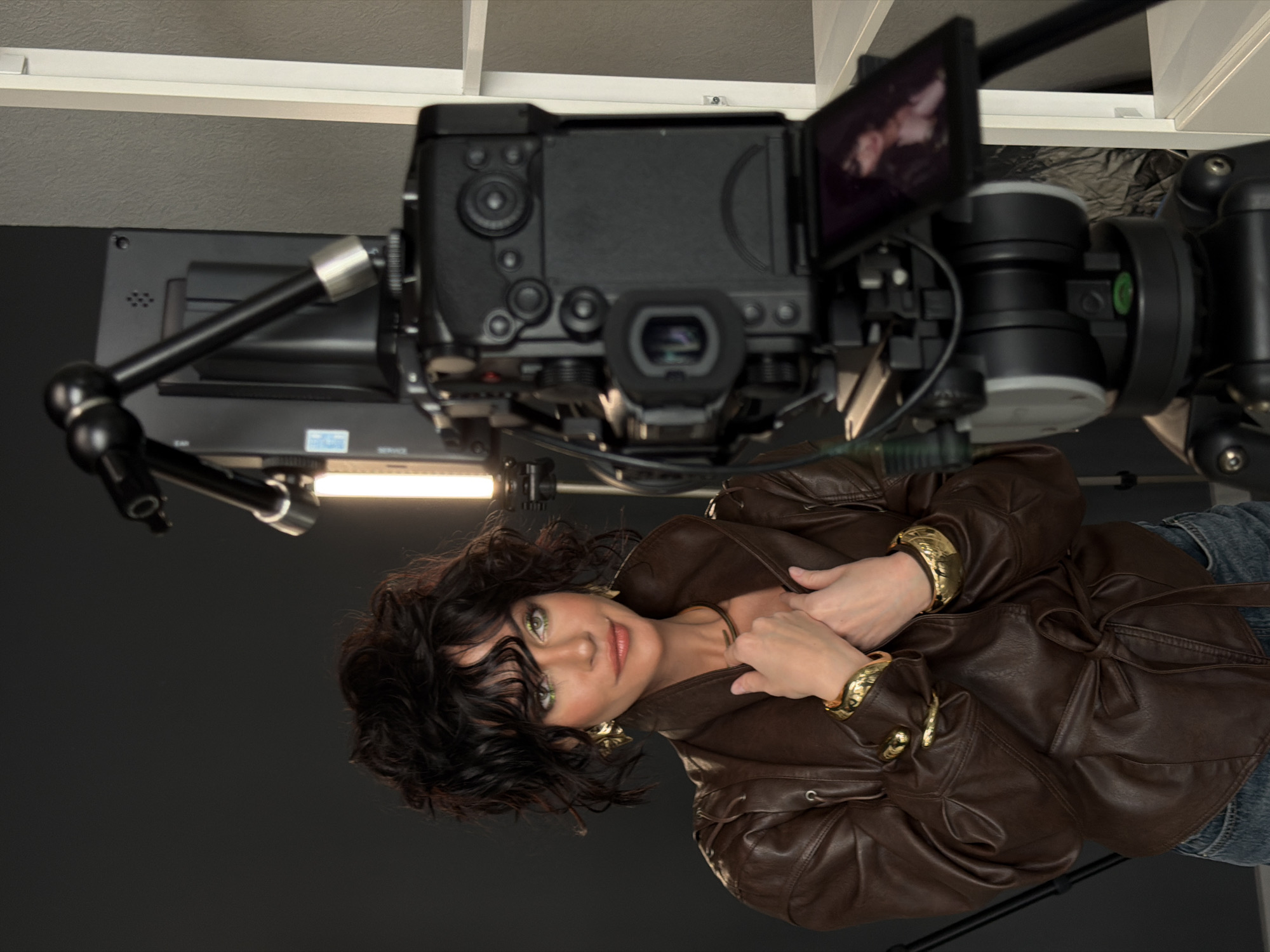 This image, currently oriented 90 degrees counter-clockwise, captures a woman during a professional photoshoot or video recording in a studio setting, contextually in Krasnoyarsk, Russia. The woman, positioned centrally, has dark, curly, shoulder-length hair and striking light-colored (possibly green or hazel) eyes, accentuated by makeup. She wears a brown leather jacket, light blue jeans, and notable gold jewelry, including large, ornate earrings, a thick choker, and two wide, textured bangles on each wrist. Her expression is a soft, confident smile as she looks towards the viewer, with her hands gently clasped over her jacket lapels.
Above and slightly behind the woman, a sophisticated camera setup is visible. This includes a professional video camera body with a large lens, mounted on a rig. An articulated external monitor, displaying a live feed of the woman, is flipped out from the camera. A bright LED light tube, attached to a flexible arm, provides illumination. The background is a plain dark gray or black wall, indicative of a controlled studio environment, with a white ceiling structure visible at the very top. Faint, blurry text "EAR" and "SERVICE" is discernable on labels attached to the camera equipment. The scene suggests a professional production, with the woman as the primary subject.