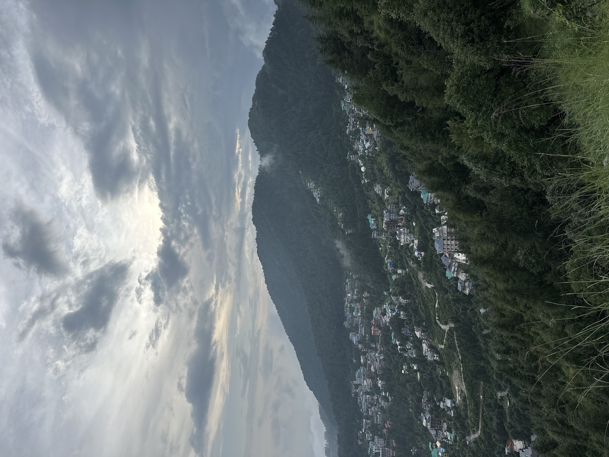 An aerial, high-angle, wide shot captures a cloudy, overcast sky over a lush, green mountainside dotted with buildings and trees in Bhagsu, India. The town sprawls across the lower slopes of a large mountain, with buildings clustered together and winding roads snaking through the landscape. Numerous trees cover the mountains, with a dense forest dominating the foreground on the right. The sky is filled with a mix of white and dark gray clouds, with some patches of lighter sky suggesting the sun may be attempting to break through. The overall impression is one of a densely populated mountain town nestled within a heavily forested natural environment under a dramatic sky.