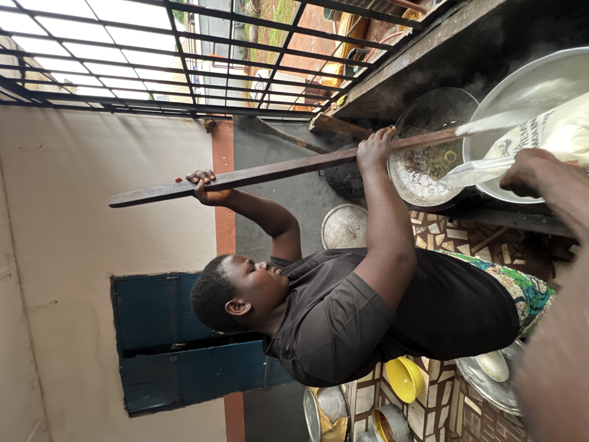 In Nkpor, Nigeria, a woman in a black t-shirt and a patterned skirt is actively engaged in cooking. She is stirring a large pot with a long wooden spoon, her focus evident on the task at hand. Beside her, another person is adding a bag of white powder, possibly flour or fufu, to a large silver bowl. The scene appears to be an outdoor kitchen, with a metal grate visible through a window, suggesting an urban setting. Various cooking utensils and pots are scattered around, indicating a busy culinary environment. The overall impression is one of domestic activity and food preparation.