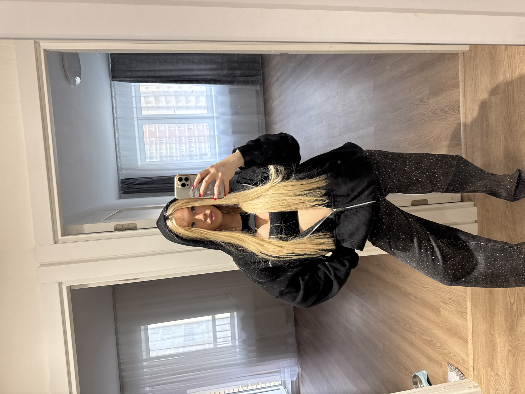 A young woman with long blonde hair takes a mirror selfie in an apartment. She is wearing a black zip-up hoodie over a black crop top and dark, embellished pants. The apartment appears to be newly renovated or unfurnished, with light wood floors and plain walls. Through the doorways, one can see windows with sheer white curtains and darker grey drapes. The buildings visible outside the windows suggest an urban environment, potentially Saint Petersburg, Russia, characterized by multi-story residential buildings. A dog bowl and a mop are visible on the floor to the left.