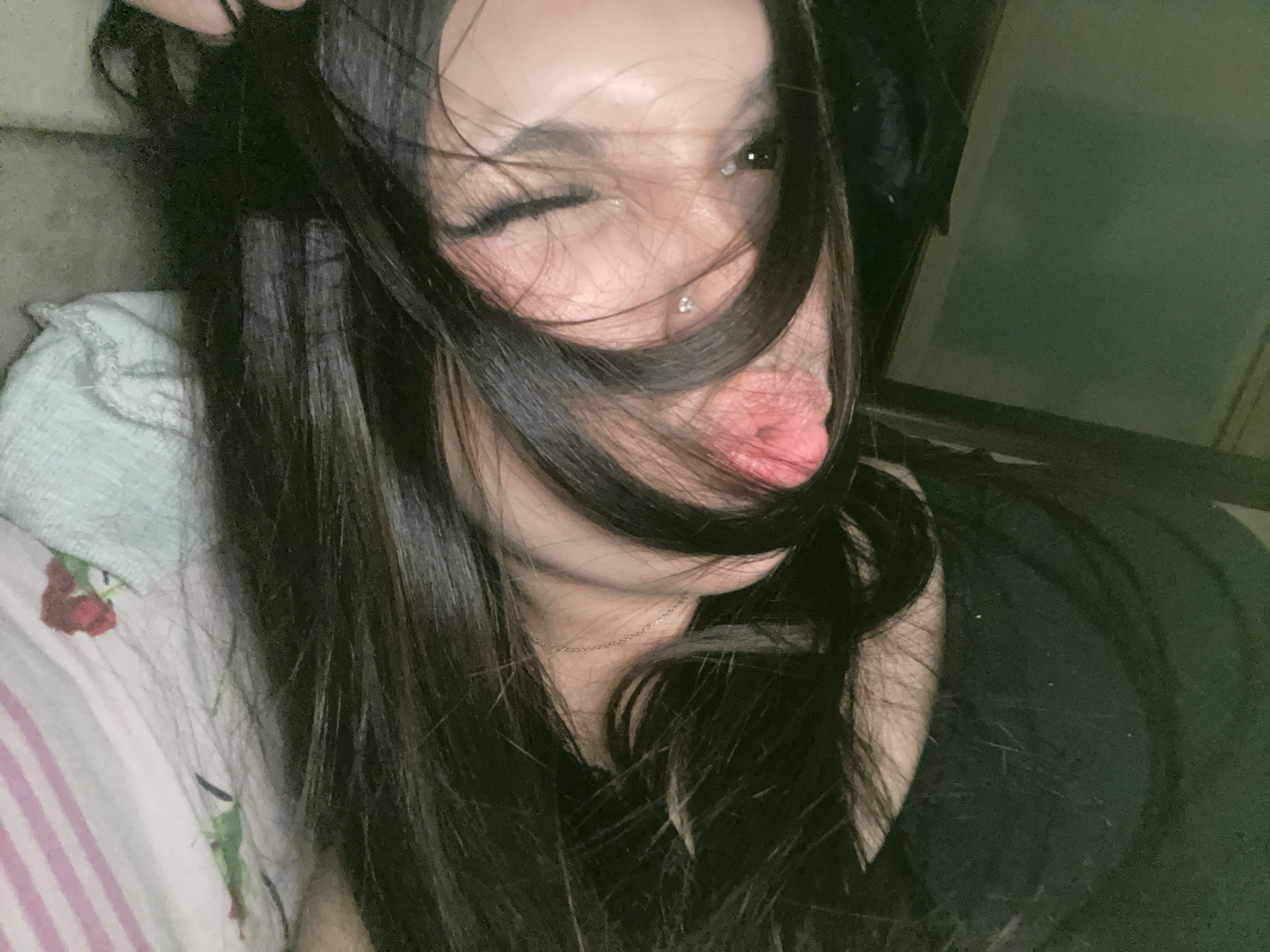 A close-up, informal portrait captures a person, likely a young woman, indoors in Saint Petersburg, Russia. Her long, dark, straight hair covers much of her face, giving a casual and intimate feel to the image. She is shown making a playful expression, winking with her right eye and sticking her tongue slightly out to the side. A small, silvery facial piercing is visible below her lower lip. Her cheeks show a soft blush, and she has visible, long eyelashes. Around her neck, a thin silver chain necklace is visible. She appears to be wearing a dark-colored top. In the lower left, a light-colored, possibly ruffled, fabric is visible, suggesting a pillow or bedding, alongside another fabric with pink stripes and a red rose pattern. The background is dark and indistinct, indicating an indoor setting with dim, artificial lighting, suggesting the photo was taken in the evening or at night. The overall mood conveyed is playful and lighthearted. There is no visible text in the image.