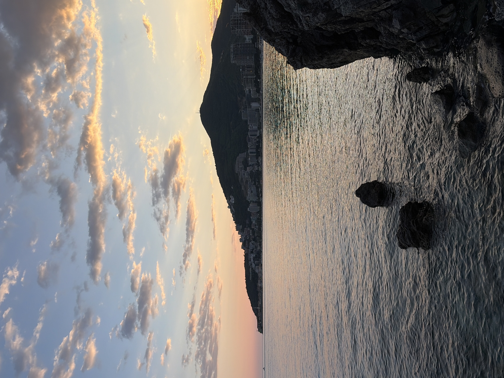 The image captures a serene coastal landscape at sunset in Budva, Montenegro. The sky, bathed in the soft, warm light of the setting sun, transitions from a bright golden glow near the horizon to a delicate blue higher up. Scattered, irregularly shaped clouds are illuminated with hues of orange, pink, and gold, adding depth and texture to the heavens.
Below the horizon line, the calm sea stretches across the midground. Its surface is subtly textured with small ripples, reflecting the ambient light and the gentle colors of the sky. Far in the distance on the water, a tiny, indistinct boat can be seen.
On the right side of the frame, a large, dark mountain or hill rises majestically from the coastline. Its slopes are dotted with numerous buildings, likely coastal resorts or residential structures, silhouetted against the bright sky and extending along the water's edge. These structures appear to be part of a developed coastal area.
In the immediate foreground, on the left, a dark, rugged rock formation or cliff face juts into the scene, creating a strong silhouette and casting deep shadows over the water nearby. A couple of dark rocks are also visible partially submerged in the water close to this formation. The overall mood is one of tranquil beauty during the golden hour. No visible text is present in the image.