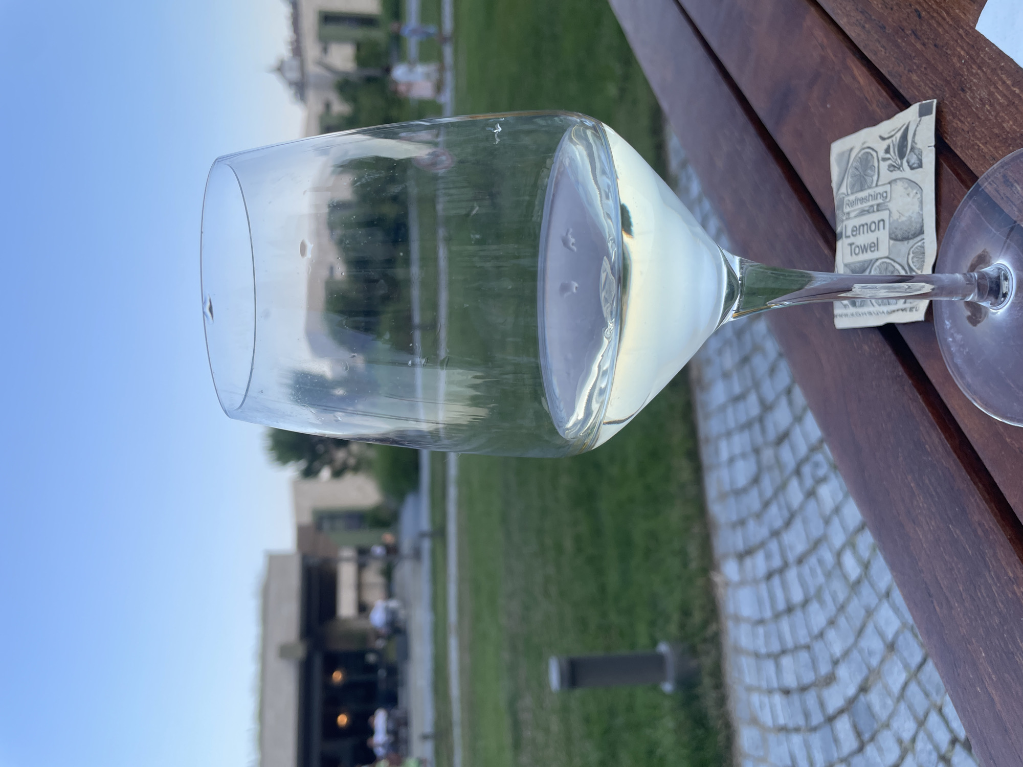 In the foreground, a stemmed glass filled with white wine sits on a wooden table. A refreshing lemon towel is placed on the table in front of the glass. In the background, a grassy area is visible, with a paved walkway and a building in the distance. The sky above is a clear blue, suggesting it is daytime. The scene appears to be an outdoor seating area of a restaurant or cafe in Obzor, Bulgaria. There are blurred figures of people in the background, some seated and others standing.