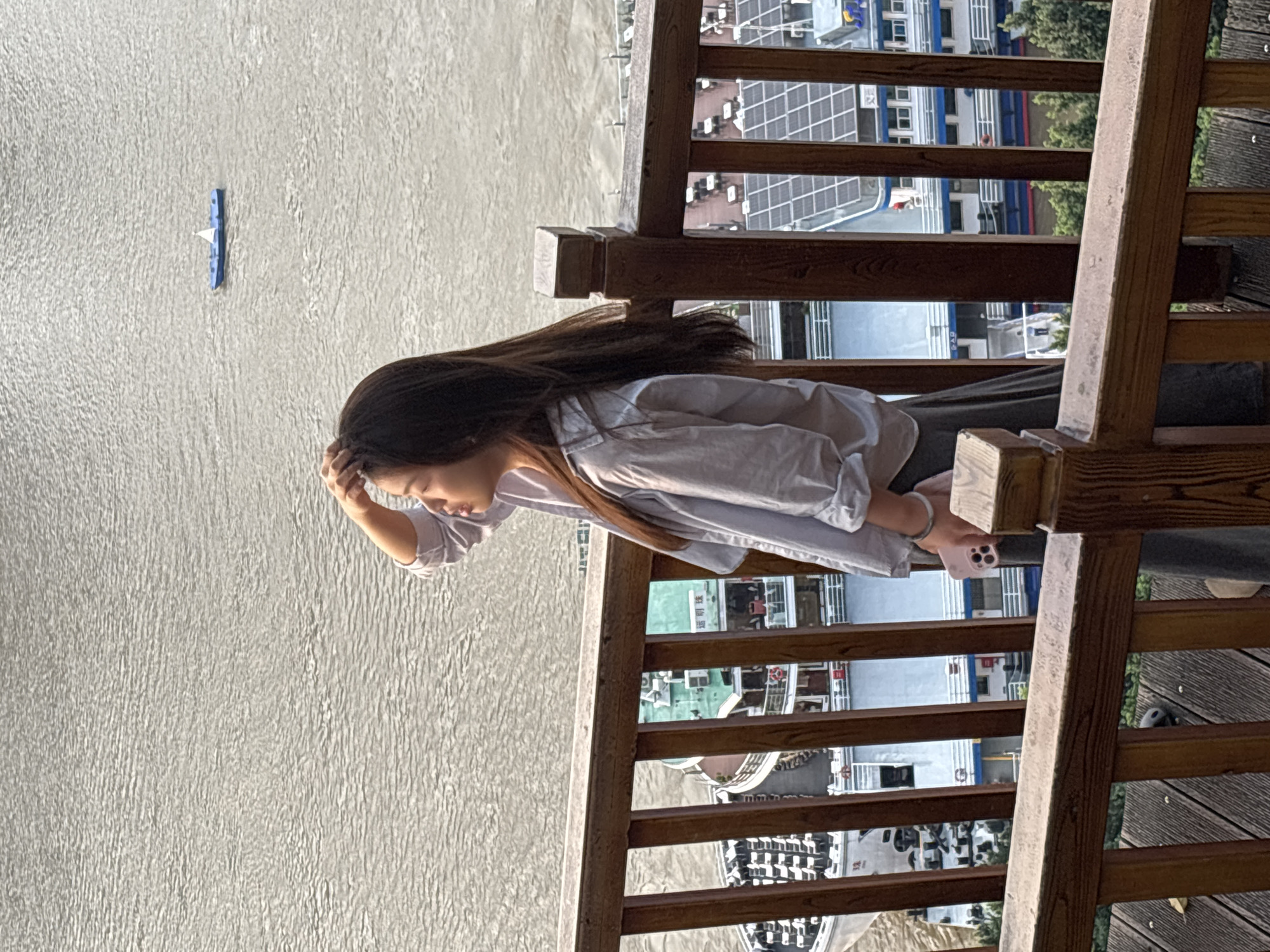 A medium shot depicts a woman standing on a wooden platform overlooking a body of water, likely the Yangtze River in Yuzhong District, Chongqing, China. She is partially visible, from the mid-thigh up, with dark hair, wearing a light-colored button-down shirt, dark pants, and holding a pink cell phone. The wooden platform has a railing, allowing a view of the river, where a small boat with a white sail is present. In the background, buildings are visible across the river, showcasing the urban landscape of Chongqing. The scene suggests an outdoor observation point in an urban environment.