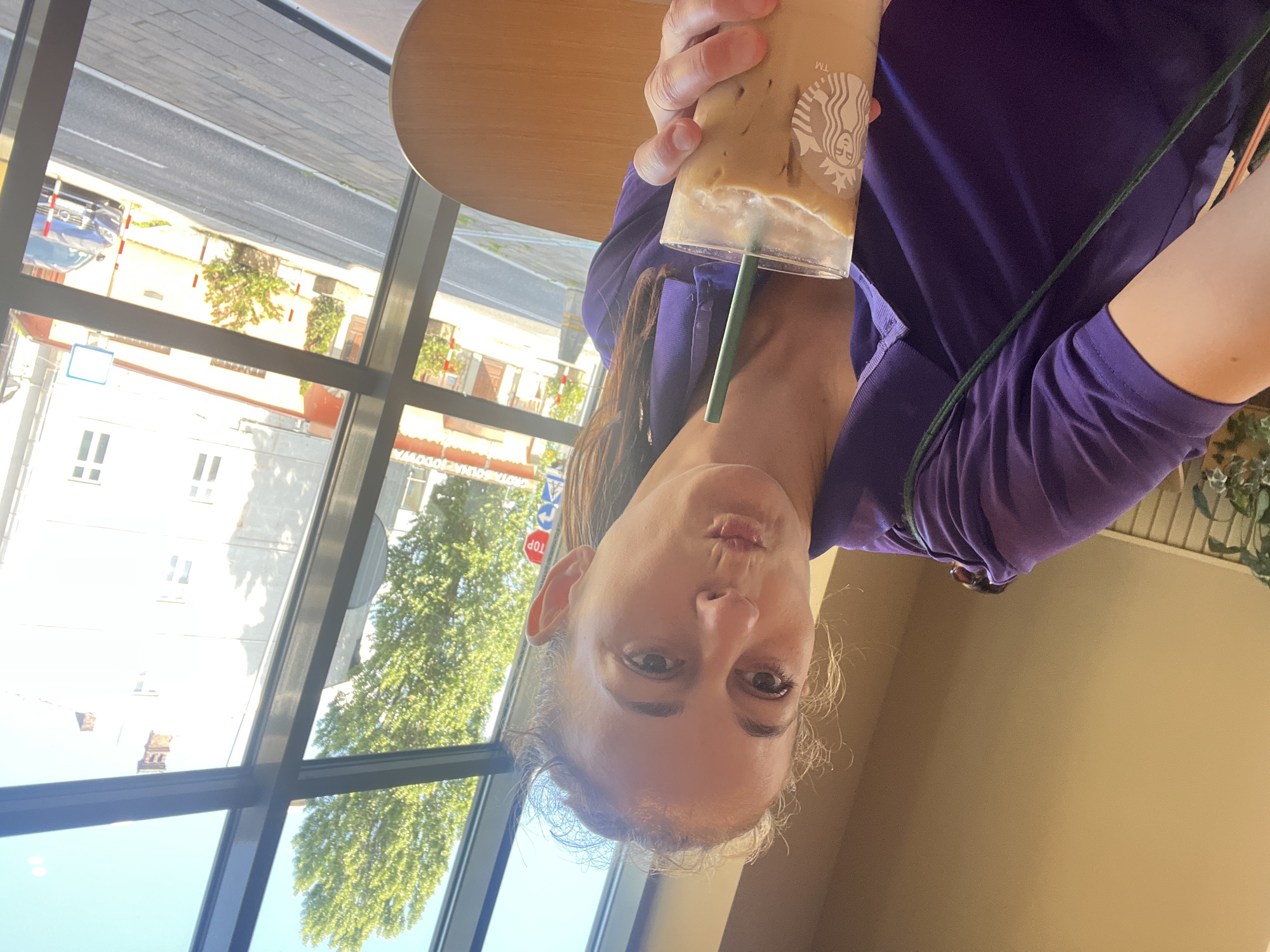 An overhead, medium shot shows a woman with blonde hair pulled back in a ponytail, wearing a purple polo shirt, holding a Starbucks iced coffee in a clear glass with a green straw. She is indoors, seated at a wooden table, with a large window behind her. Outside the window, a tree with green leaves and a light blue sky are visible. In the background, there are buildings, a street with a blue car, and a stop sign. The woman is looking at the camera with her lips puckered, as if about to take a sip of her drink.
