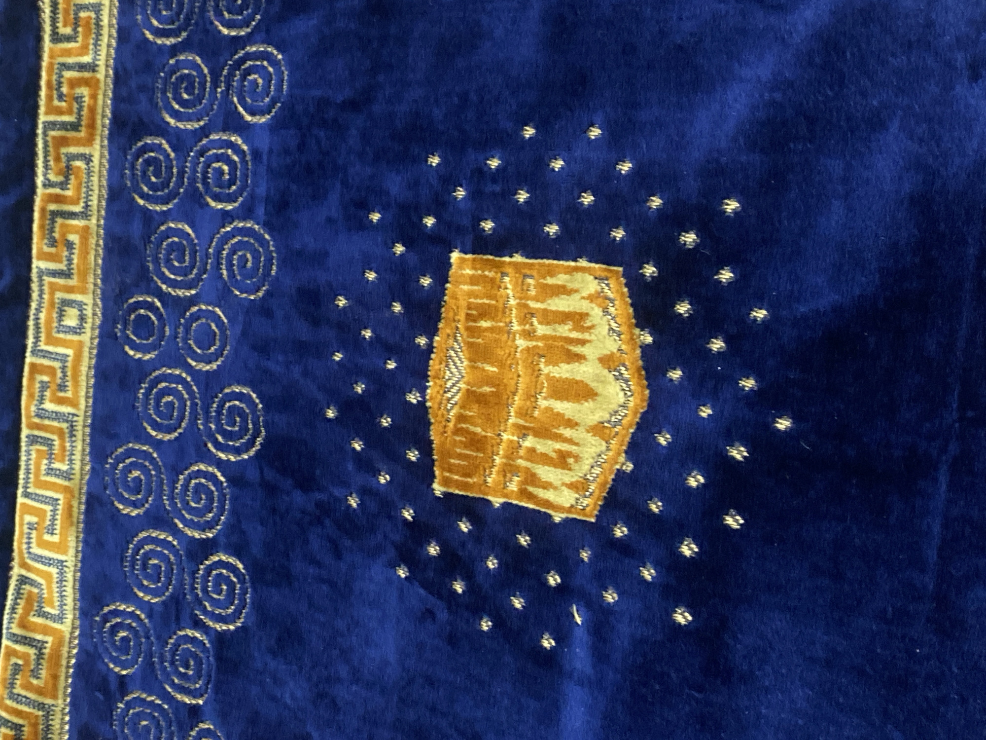 The image shows a close-up view of a prayer rug with intricate designs. The rug is a deep blue color and features a decorative border at the top with a geometric pattern in yellow and white. Below the border, there are rows of gold spiral designs. In the center of the rug, there is an embroidered image of the Kaaba in Mecca, also in gold and white, surrounded by small gold star-like embellishments. The texture of the rug appears to be soft, possibly velvet. The image does not contain any information about the city of Banjarbaru, Indonesia, or any people, actions, or interactions.