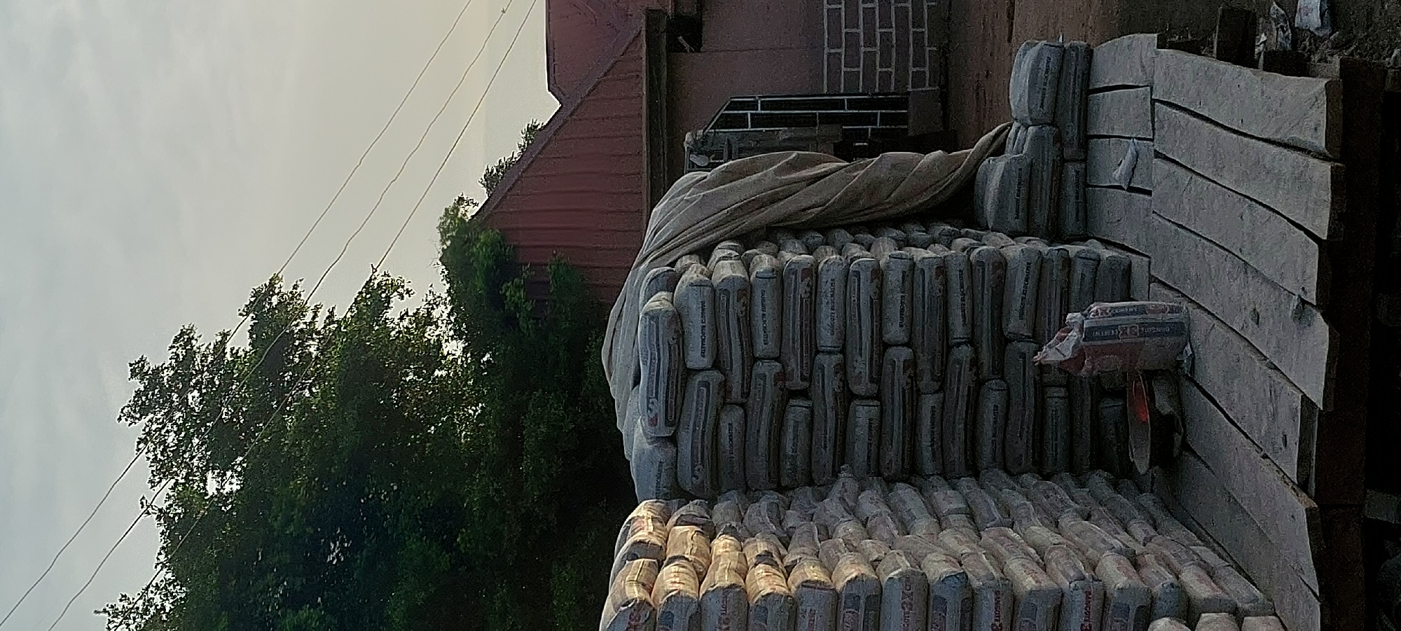 In Jalingo, Nigeria, a construction site is depicted with stacks of bagged cement placed on wooden planks. The cement bags are predominantly gray with some branding visible. A tarp partially covers one of the cement stacks. In the background, a building with a red metal roof and brickwork is present, along with lush green trees and overhead power lines against a pale sky. A small red bowl and a single bag of cement are also visible on the wooden planks. The scene appears to be set outdoors during daylight.