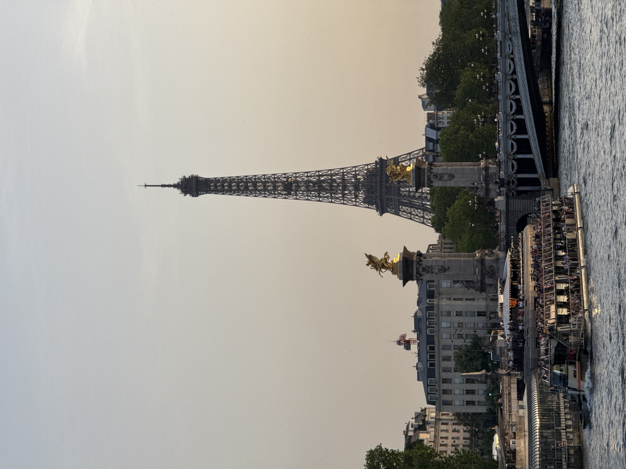 The image shows the Eiffel Tower in Paris, France, viewed from a low angle, likely from the Seine River. In the foreground, a tour boat filled with people is visible on the river. The Pont Alexandre III bridge and its gilded statues are also prominent features in the midground. The sky is overcast and hazy. Buildings along the riverbank are also visible.