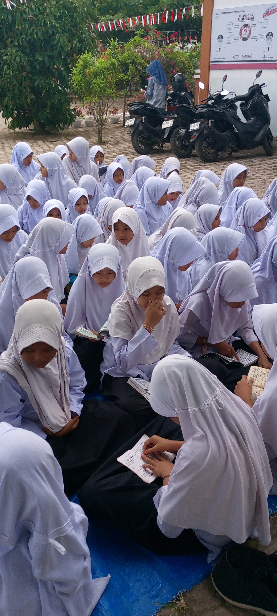 In the city of Baratpayaitek, Indonesia, a group of young girls in white hijabs and uniforms are seated on a blue tarp on the ground. The girls are engaged in various activities, with some reading from books, others looking down at papers, and one girl appears to be writing or drawing on a small notebook. The scene is outdoors, with trees and foliage in the background. Several motorcycles are parked behind the girls, and a banner with text and graphics is visible on a wall in the distance. The overall impression is one of a school or religious gathering in an outdoor setting.