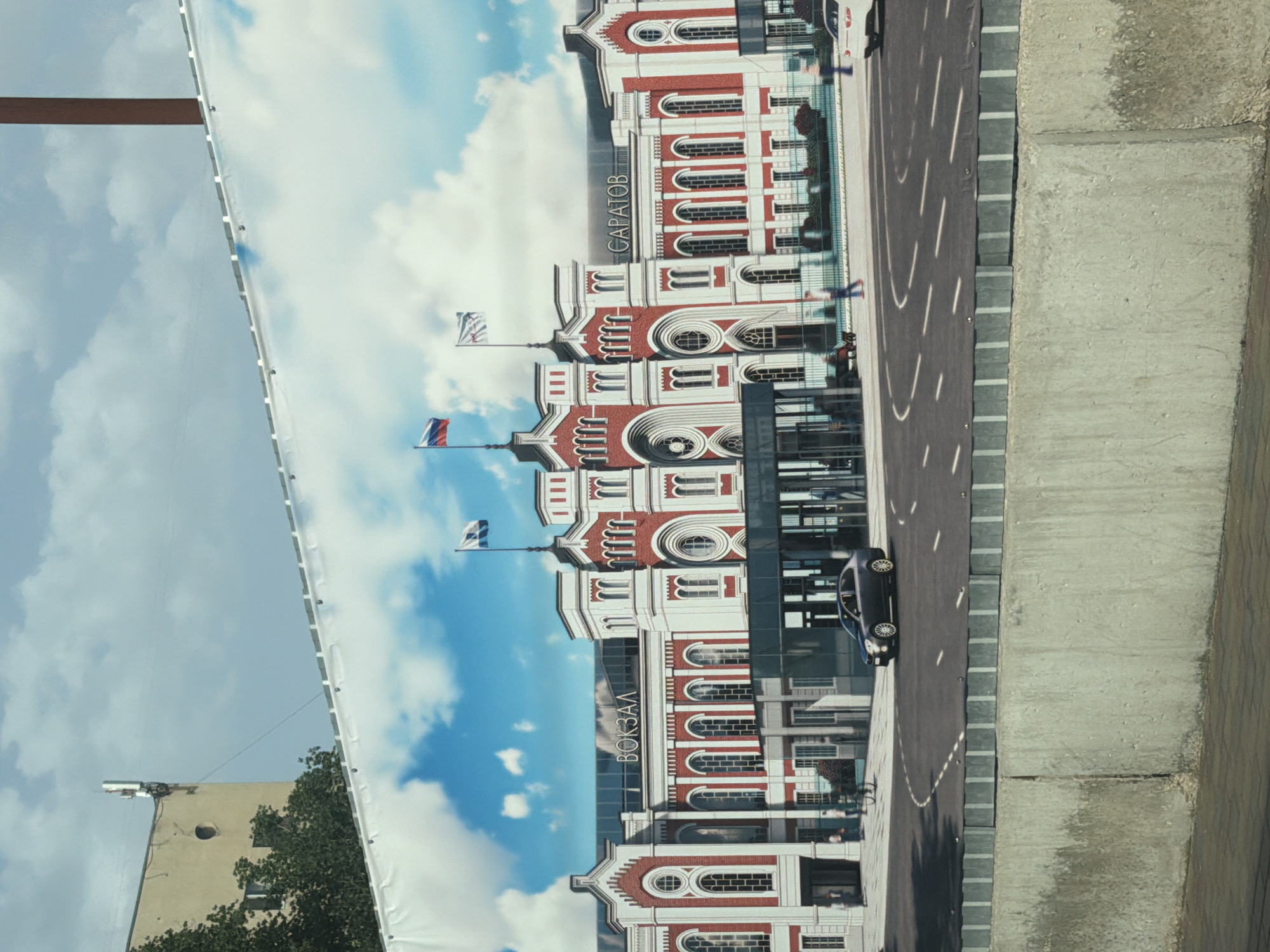 The image shows a poster of the Saratov Train Station in Russia. The building is brick-red and white, with the Cyrillic words "ВОКЗАЛ" and "САРАТОВ" vertically displayed, meaning "Train Station" and "Saratov" respectively. The sky in the background is blue with white clouds. A road is depicted, with a black car parked near the train station building. A curb separates the road and the sidewalk. The poster is attached on the left and is in front of a gray building and tree with green leaves. Flags appear on the building in the poster.